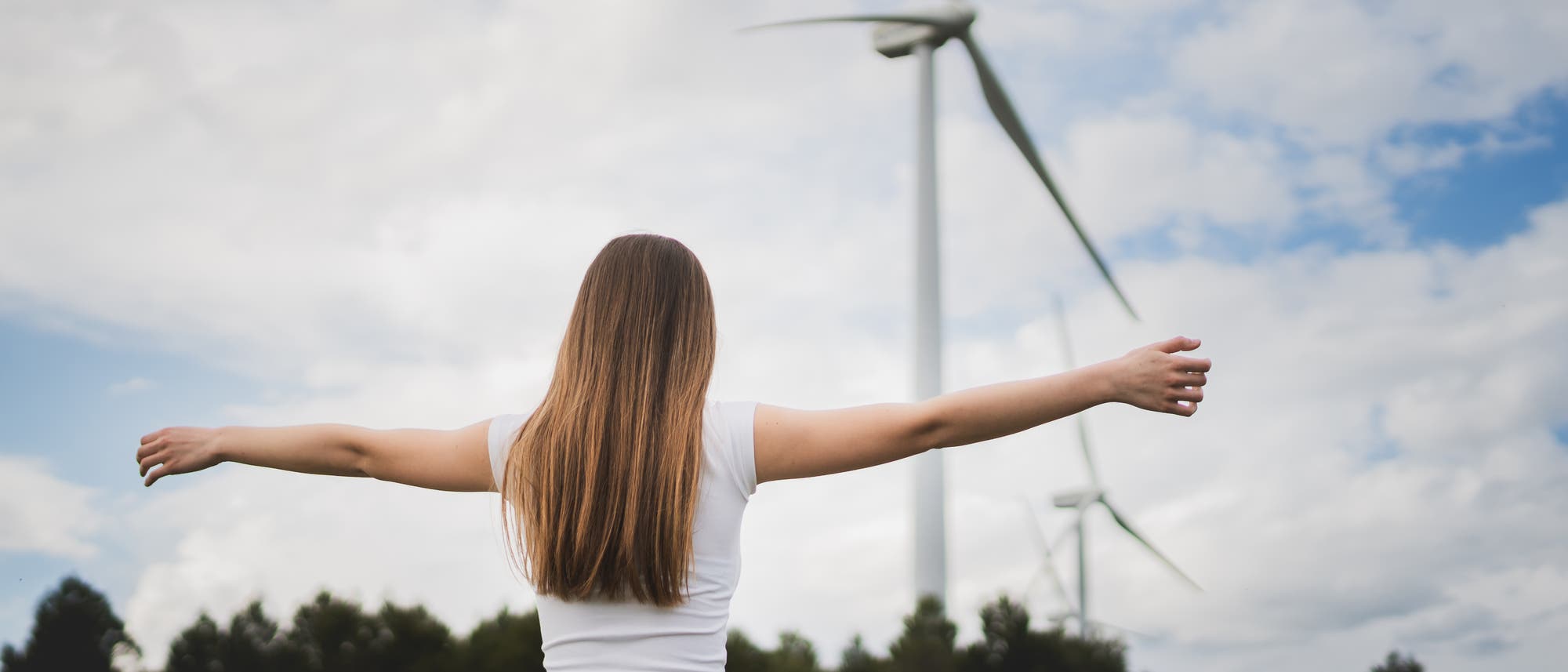 Eine Person steht mit ausgebreiteten Armen auf einer Wiese und blickt auf zwei gro&szlig;e Windkraftanlagen im Hintergrund. Der Himmel ist bew&ouml;lkt, und im Vordergrund sind B&auml;ume zu sehen. Die Szene vermittelt ein Gef&uuml;hl von Freiheit und Nachhaltigkeit.