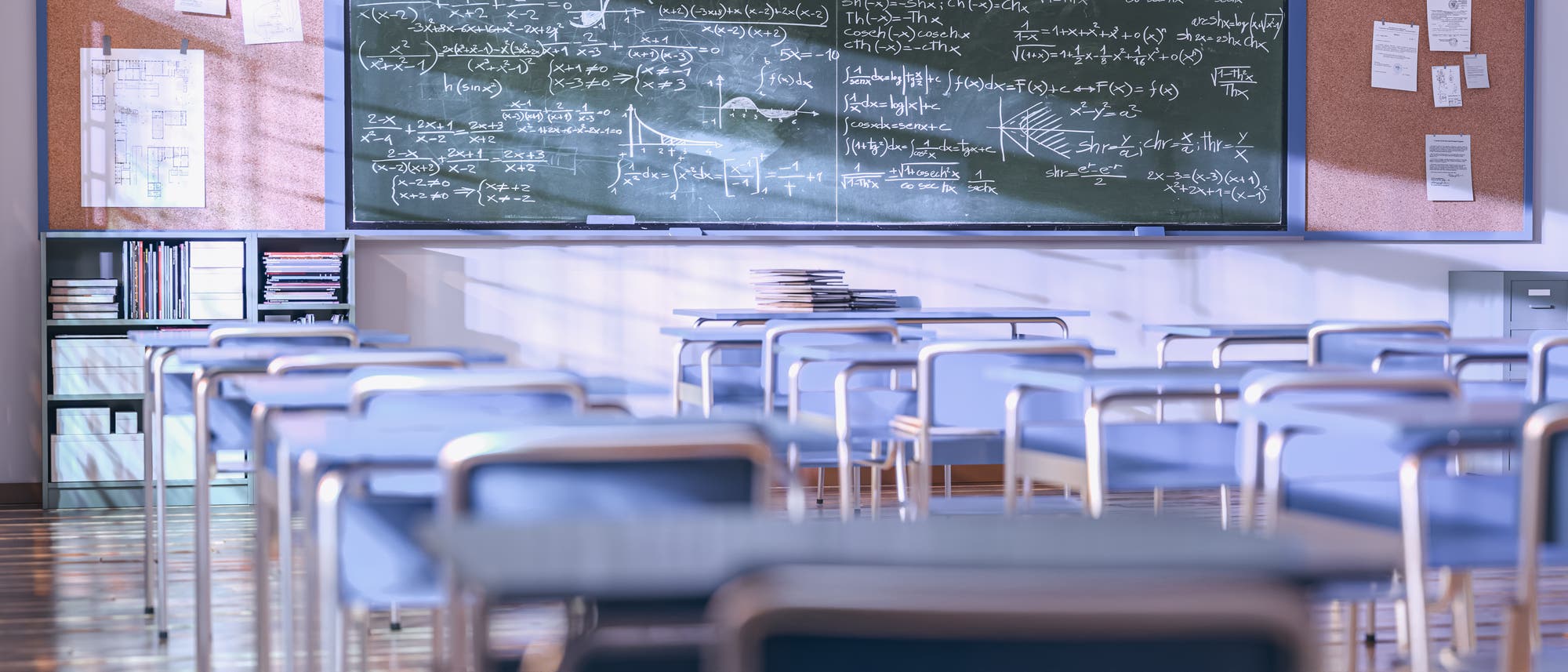 Ein leeres Klassenzimmer mit mehreren Reihen von blauen St&uuml;hlen und Tischen. Im Hintergrund befindet sich eine gr&uuml;ne Tafel, die mit komplexen mathematischen Formeln und Diagrammen beschrieben ist. An der Wand neben der Tafel h&auml;ngen Korktafeln mit Notizen und Zeichnungen. Der Raum ist gut beleuchtet, und auf einigen Tischen liegen gestapelte B&uuml;cher.