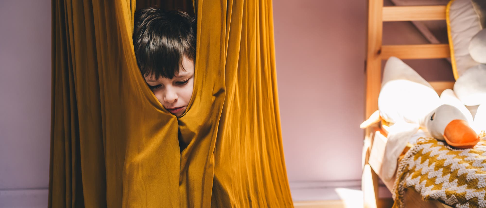 Ein Kind versteckt sich in einem senfgelben Vorhang in einem gem&uuml;tlichen Kinderzimmer. Rechts im Bild steht ein Holzbett mit einer gestrickten Decke und einem Pl&uuml;schspielzeug in Form einer Ente. Die Szene wirkt ruhig und einladend.