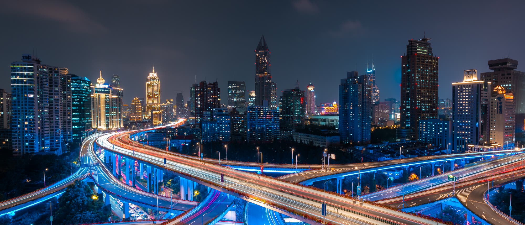 N&auml;chtliche Stadtansicht mit beleuchteten Hochh&auml;usern und einem komplexen Autobahnkreuz im Vordergrund. Die Lichter der Fahrzeuge erzeugen farbige Lichtspuren, die die Dynamik des Verkehrs verdeutlichen. Der Himmel ist dunkel, was die st&auml;dtische Beleuchtung hervorhebt.