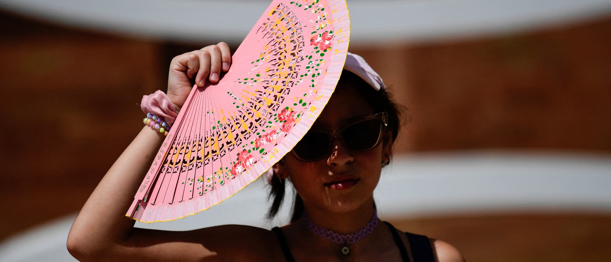 Eine Person hält einen rosa Fächer mit floralen Mustern über ihrem Kopf, um sich vor der Sonne zu schützen. Sie trägt eine Sonnenbrille und ein schwarzes Oberteil. Der Hintergrund ist unscharf und zeigt eine braune Wand mit weißen Bögen. Eine Person hält einen rosa Fächer mit floralen Mustern über ihrem Kopf, um sich vor der Sonne zu schützen. Sie trägt eine Sonnenbrille und ein schwarzes Oberteil. Der Hintergrund ist unscharf und zeigt eine braune Wand mit weißen Bögen.