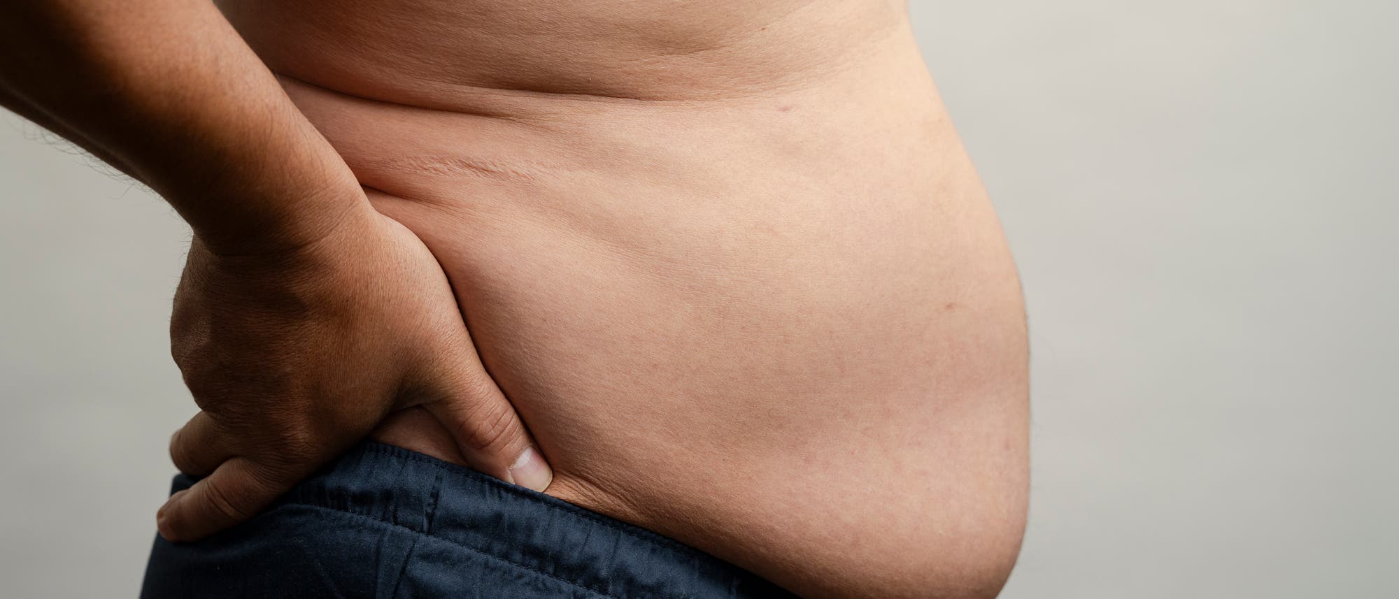 Eine Person steht seitlich und h&auml;lt eine Hand in die H&uuml;fte. Der Fokus liegt auf dem Bauchbereich. Die Person tr&auml;gt eine dunkle Hose. Der Hintergrund ist neutral und hell.