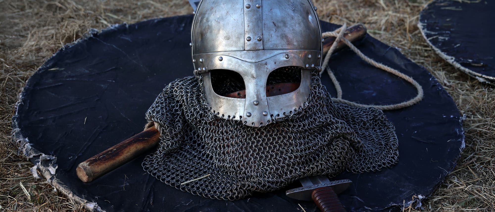 Ein mittelalterlicher Helm aus Metall liegt auf einem runden Schild, umgeben von einer Kettenhaube. Daneben befinden sich ein Holzgriff und ein Schwert. Der Hintergrund besteht aus trockenem Gras.