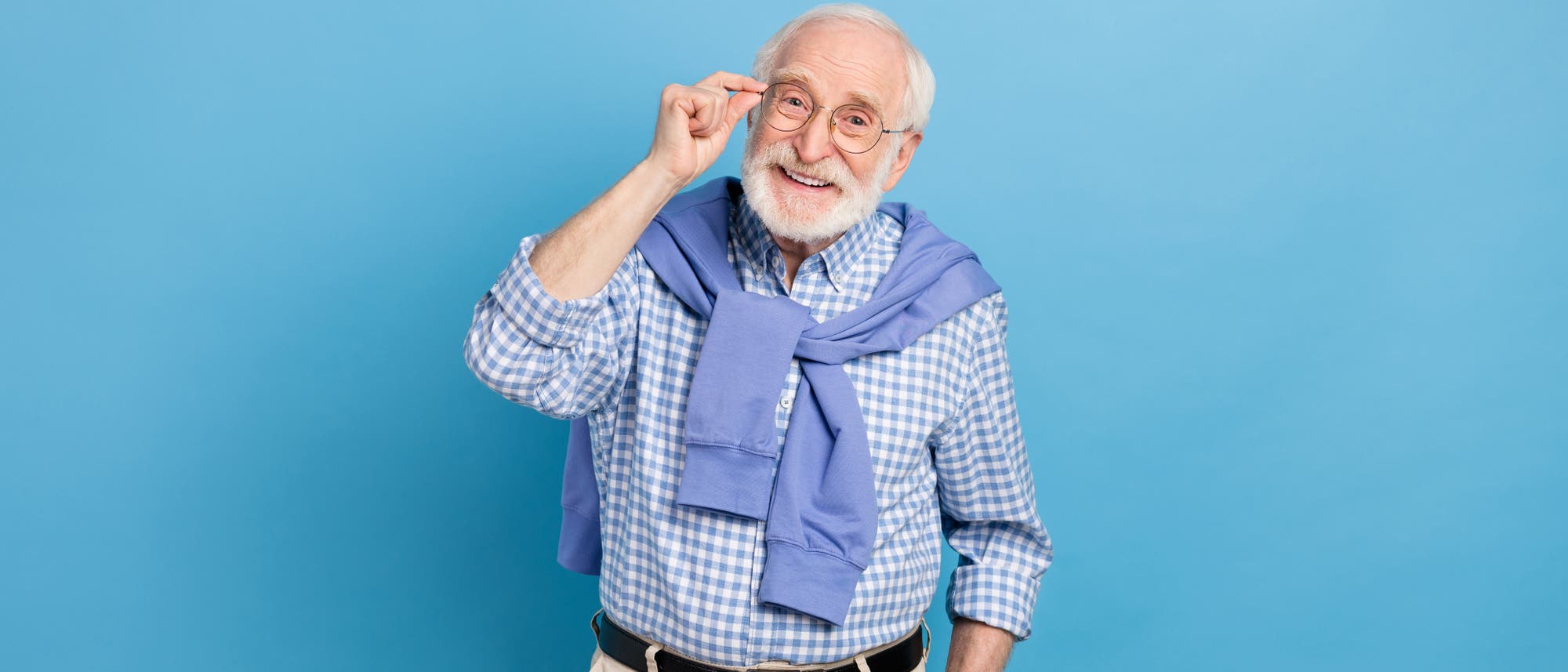 Ein älterer Mann mit Brille lächelt freundlich in die Kamera. Er trägt ein kariertes Hemd und hat einen Pullover um die Schultern gebunden. Der Hintergrund ist einfarbig blau. Ein älterer Mann mit Brille lächelt freundlich in die Kamera. Er trägt ein kariertes Hemd und hat einen Pullover um die Schultern gebunden. Der Hintergrund ist einfarbig blau.