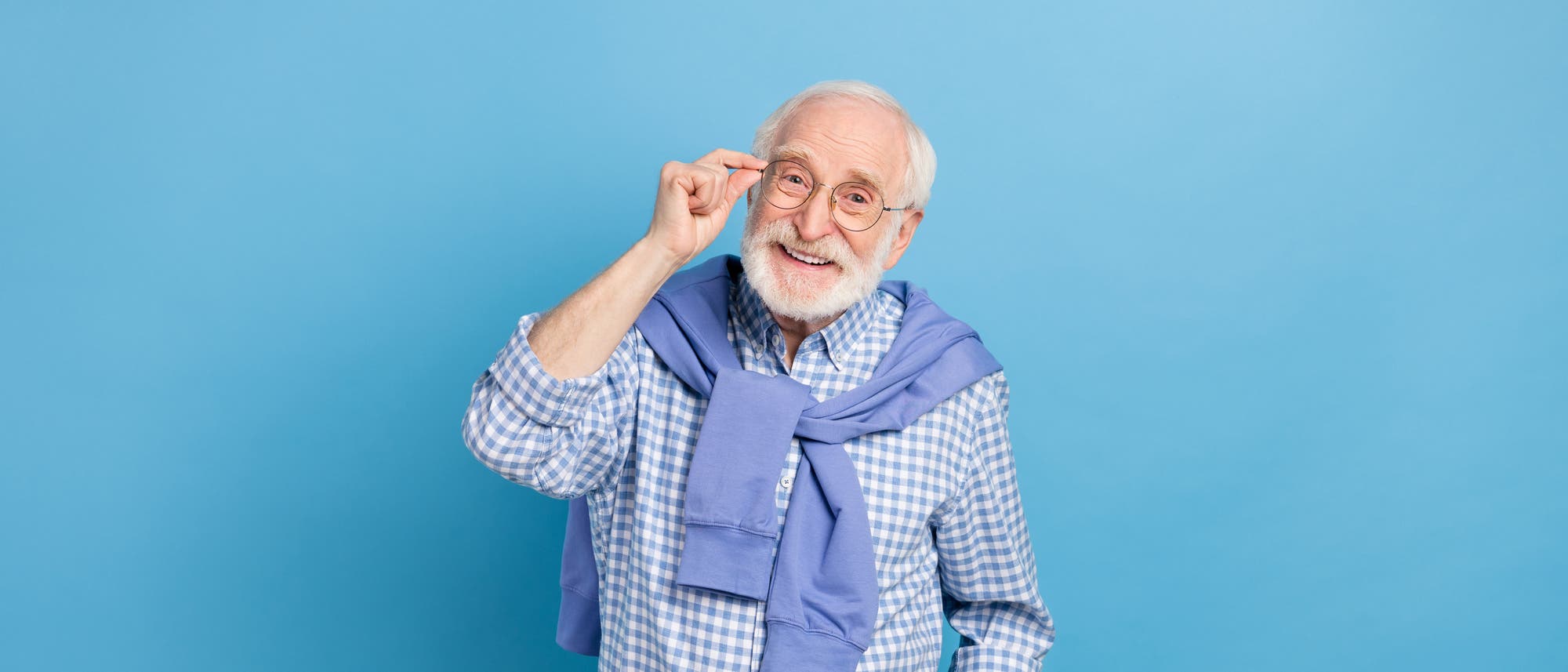 Ein älterer Mann mit Brille lächelt freundlich in die Kamera. Er trägt ein kariertes Hemd und hat einen Pullover um die Schultern gebunden. Der Hintergrund ist einfarbig blau. Ein älterer Mann mit Brille lächelt freundlich in die Kamera. Er trägt ein kariertes Hemd und hat einen Pullover um die Schultern gebunden. Der Hintergrund ist einfarbig blau.
