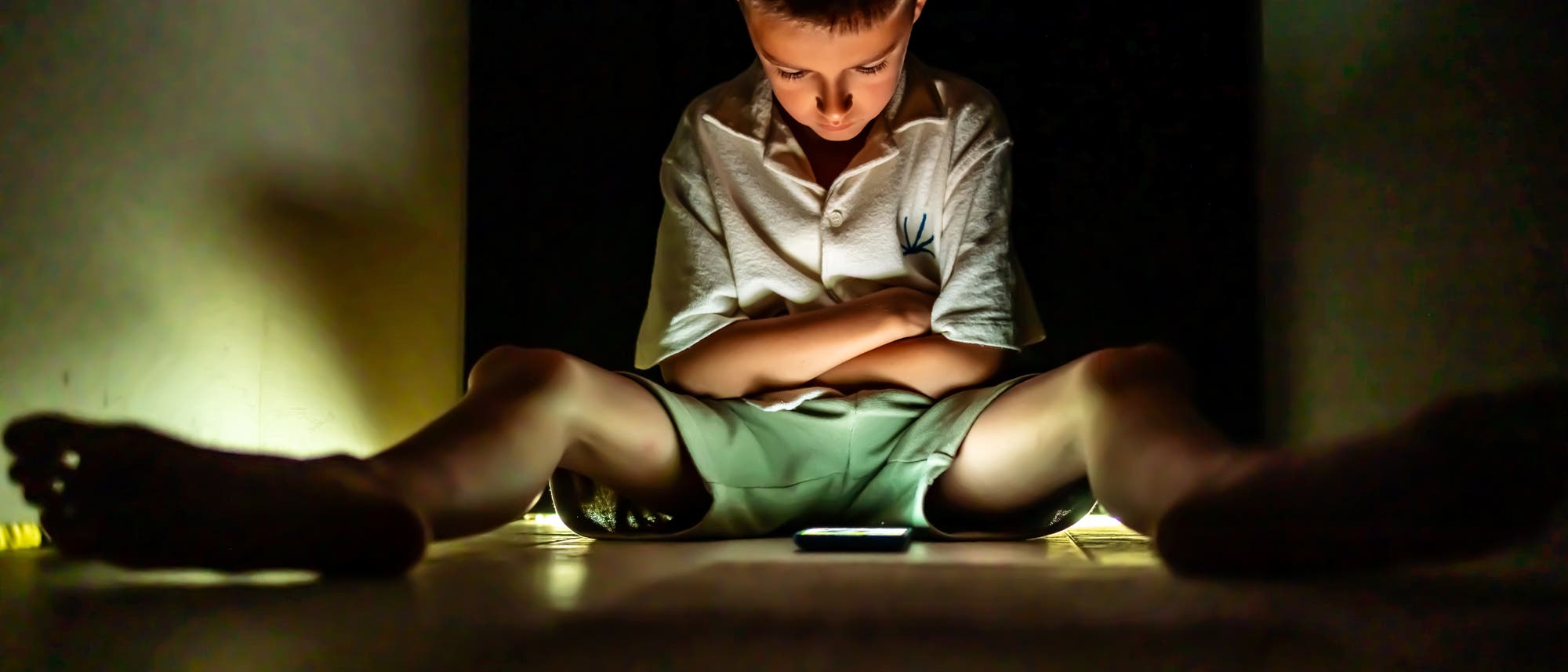 Ein Kind sitzt mit verschr&auml;nkten Armen auf dem Boden in einem dunklen Raum, beleuchtet von einem Smartphone, das vor ihm liegt. Die Szene vermittelt eine nachdenkliche oder konzentrierte Stimmung.