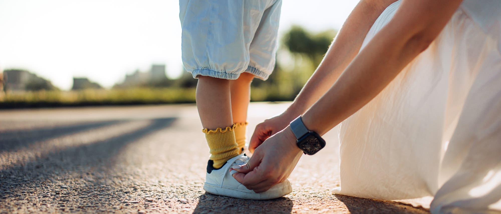 Eine erwachsene Person kniet auf einer Straße und bindet einem Kind die Schnürsenkel. Das Kind trägt ein gestreiftes Oberteil, kurze Hosen und gelbe Socken. Die Szene findet bei Tageslicht statt, und im Hintergrund sind Bäume und ein unscharfer Horizont zu sehen. Eine erwachsene Person kniet auf einer Straße und bindet einem Kind die Schnürsenkel. Das Kind trägt ein gestreiftes Oberteil, kurze Hosen und gelbe Socken. Die Szene findet bei Tageslicht statt, und im Hintergrund sind Bäume und ein unscharfer Horizont zu sehen.