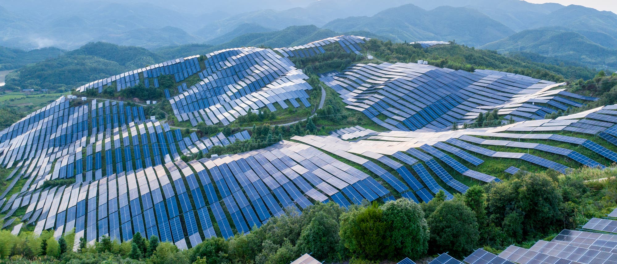 Luftaufnahme eines gro&szlig;en Solarparks in h&uuml;geliger Landschaft. Reihen von Solarpaneelen erstrecken sich &uuml;ber die gr&uuml;nen H&uuml;gel, w&auml;hrend im Hintergrund eine Bergkette unter einem klaren Himmel zu sehen ist.