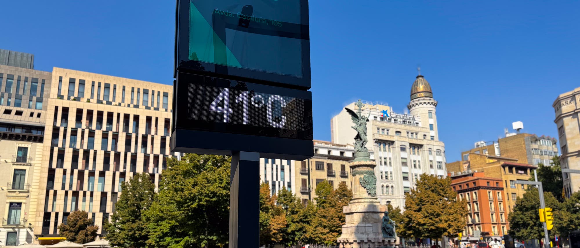 Eine städtische Szene mit einem digitalen Thermometer im Vordergrund, das 41°C anzeigt. Im Hintergrund sind moderne und historische Gebäude sowie ein Brunnen mit einer Statue zu sehen. Der Himmel ist klar und blau, was auf einen sonnigen Tag hinweist. 