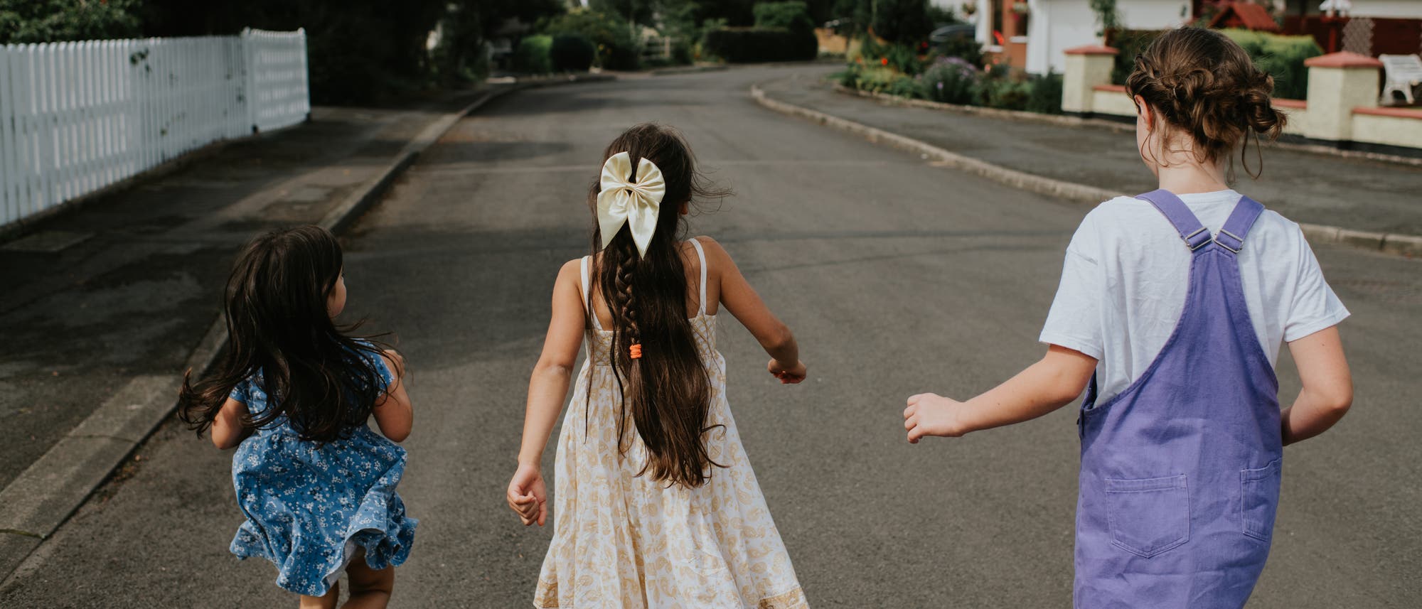 Drei Kinder laufen eine ruhige Wohnstra&szlig;e entlang. Sie tragen Sommerkleidung, darunter Kleider und ein Latzkleid. Die Umgebung ist von H&auml;usern und G&auml;rten ges&auml;umt, und die Stra&szlig;e ist leer. Ein Kind hat eine gro&szlig;e Schleife im Haar. Die Szene vermittelt ein Gef&uuml;hl von Unbeschwertheit und Sommerfreude.