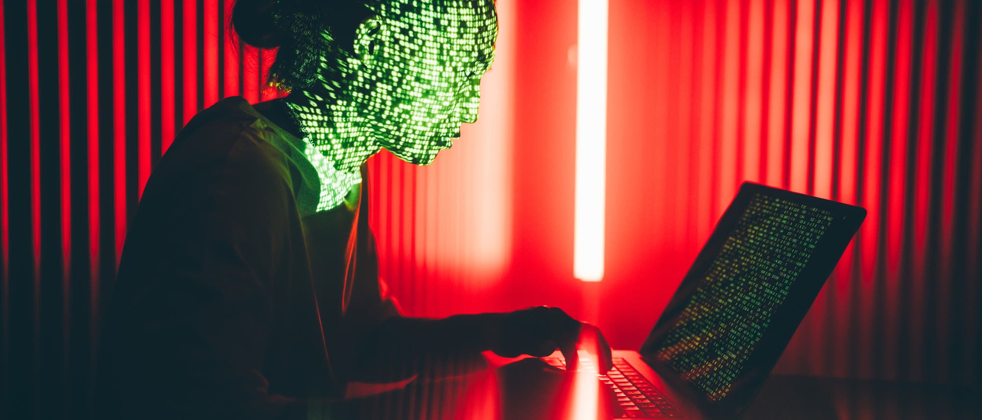 Eine Person sitzt vor einem Laptop in einem dunklen Raum, der von rotem Licht beleuchtet wird. Das Gesicht der Person ist mit gr&uuml;nem, digitalem Code &uuml;berlagert, was auf Themen wie Technologie oder Cyberwelt hinweist. Der Hintergrund zeigt vertikale Linien und eine leuchtende Lichtquelle.