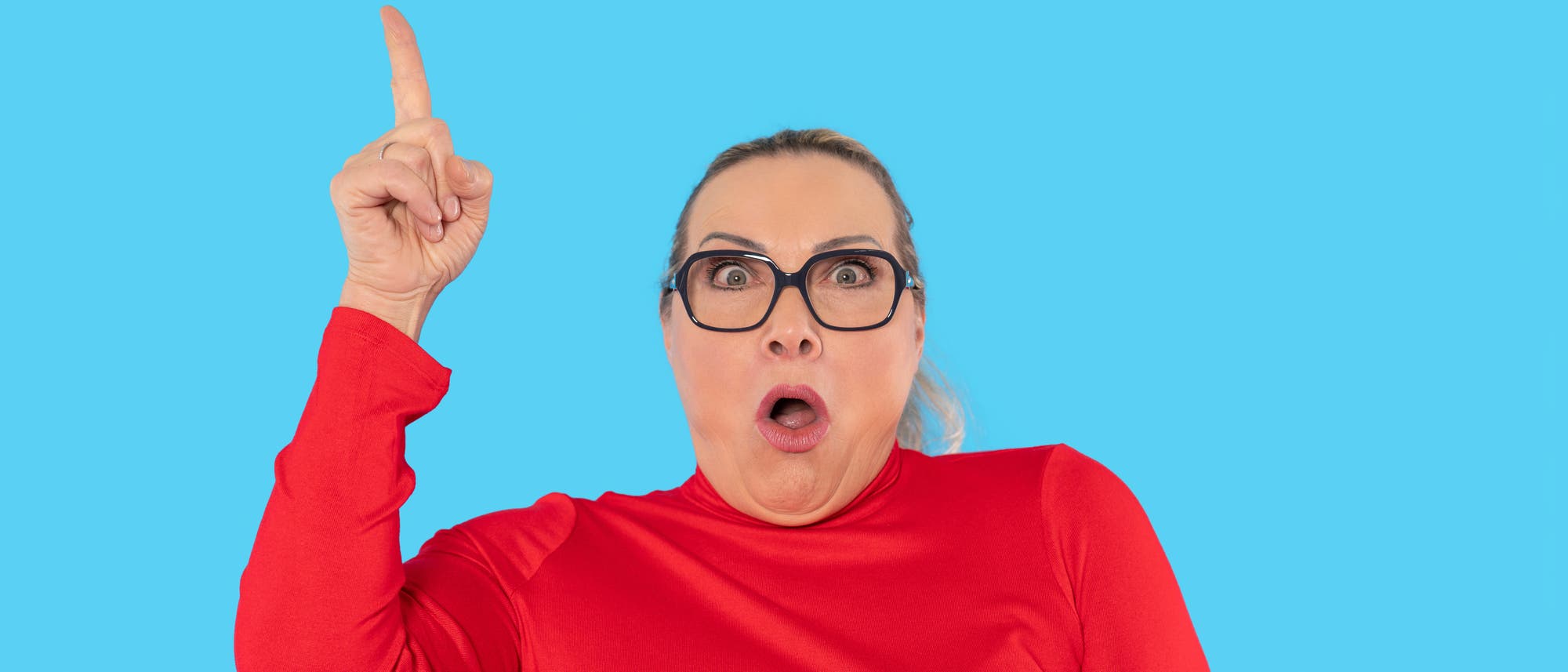 Eine Person in einem roten Oberteil zeigt mit erhobenem Zeigefinger nach oben und hat einen überraschten Gesichtsausdruck. Der Hintergrund ist einfarbig blau. Eine Person in einem roten Oberteil zeigt mit erhobenem Zeigefinger nach oben und hat einen überraschten Gesichtsausdruck. Der Hintergrund ist einfarbig blau.