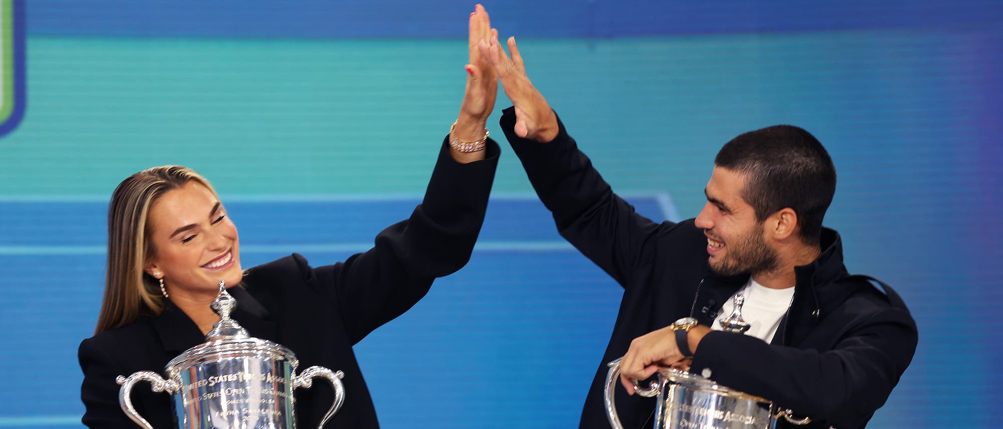 Zwei Personen sitzen nebeneinander und geben sich ein High-Five. Beide halten jeweils einen großen silbernen Pokal in der anderen Hand. Die Szene vermittelt Freude und Erfolg. Der Hintergrund ist blau.