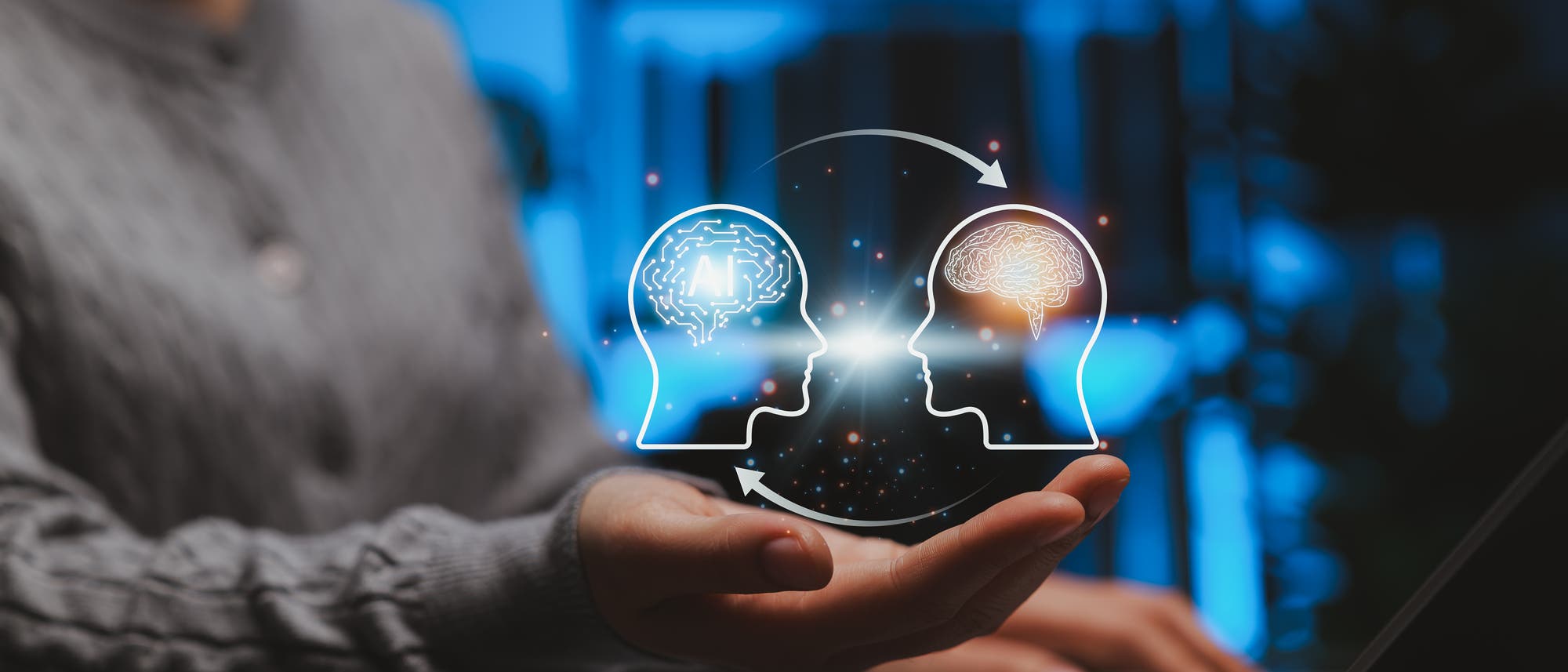 Eine Person h&auml;lt eine holografische Darstellung von zwei K&ouml;pfen &uuml;ber einem Laptop. Der linke Kopf zeigt ein Schaltkreis-Muster mit den Buchstaben "AI", w&auml;hrend der rechte Kopf ein Gehirn darstellt. Pfeile verbinden die beiden K&ouml;pfe und symbolisieren den Austausch zwischen k&uuml;nstlicher Intelligenz und menschlichem Denken. Der Hintergrund ist in Blau- und Graut&ouml;nen gehalten, was eine futuristische Atmosph&auml;re schafft.