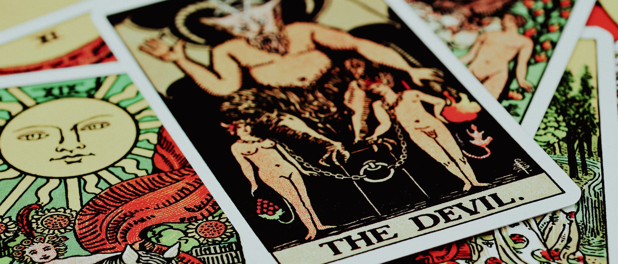 Ein Stapel Tarotkarten, im Vordergrund die Karte "The Devil". Die Karte zeigt eine mythologische Figur mit Hörnern, die über zwei angeketteten Menschen thront. Um die Hauptkarte herum sind weitere Tarotkarten mit verschiedenen Symbolen und Illustrationen sichtbar, darunter eine Karte mit einer Sonne. Die Farben sind lebendig und kontrastreich, was den mystischen und symbolischen Charakter der Karten betont. Ein Stapel Tarotkarten, im Vordergrund die Karte "The Devil". Die Karte zeigt eine mythologische Figur mit Hörnern, die über zwei angeketteten Menschen thront. Um die Hauptkarte herum sind weitere Tarotkarten mit verschiedenen Symbolen und Illustrationen sichtbar, darunter eine Karte mit einer Sonne. Die Farben sind lebendig und kontrastreich, was den mystischen und symbolischen Charakter der Karten betont.