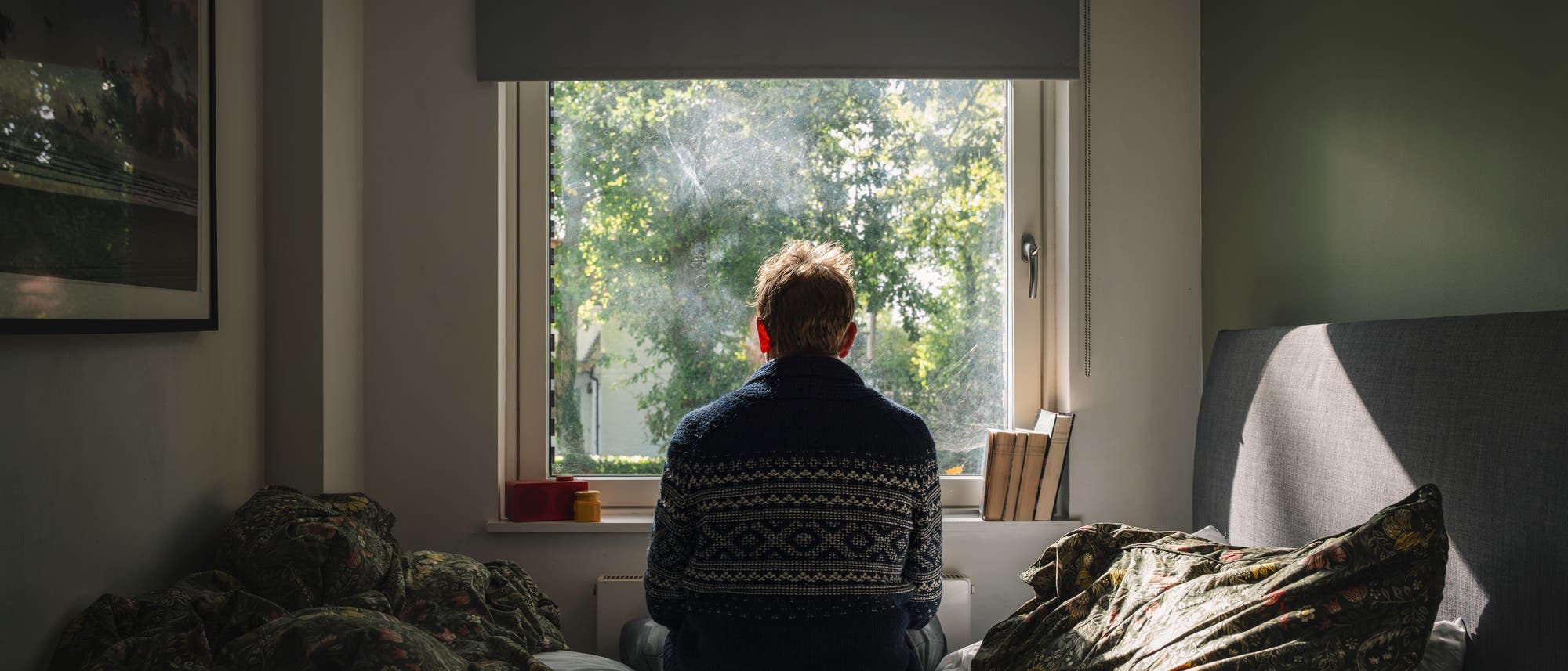Eine Person sitzt auf einem Bett und blickt aus einem Fenster, durch das Sonnenlicht auf die Szene f&auml;llt. Der Raum ist schlicht eingerichtet, mit einem Bild an der Wand und B&uuml;chern auf der Fensterbank. Drau&szlig;en sind gr&uuml;ne B&auml;ume zu sehen. Die Atmosph&auml;re wirkt ruhig und nachdenklich.