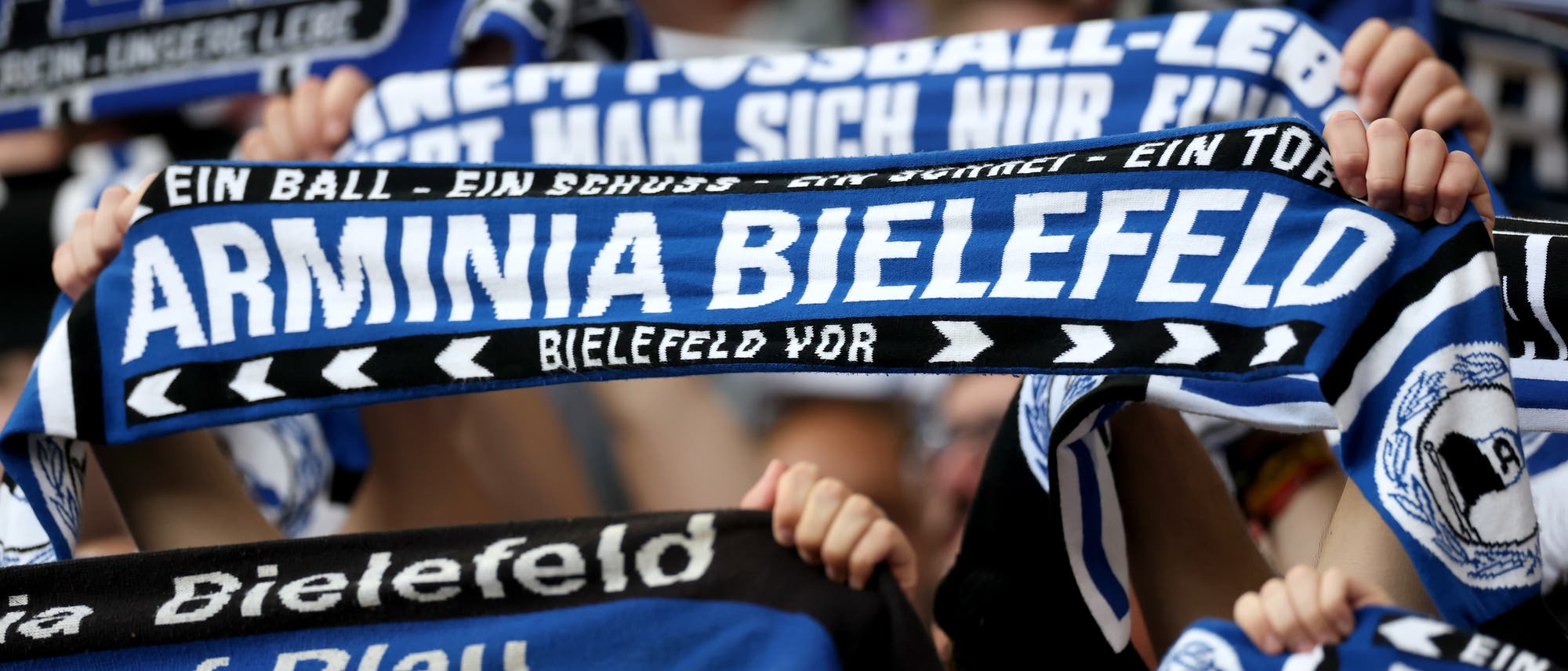 Eine Gruppe von Menschen h&auml;lt blau-wei&szlig;e Fanschals hoch, auf denen "Arminia Bielefeld" steht. Die Schals sind in verschiedenen Designs gestaltet, einige mit zus&auml;tzlichen Texten wie "Ein Ball - Ein Schuss - Ein Tor". Die Szene vermittelt eine lebhafte und unterst&uuml;tzende Atmosph&auml;re bei einem Fu&szlig;ballspiel.