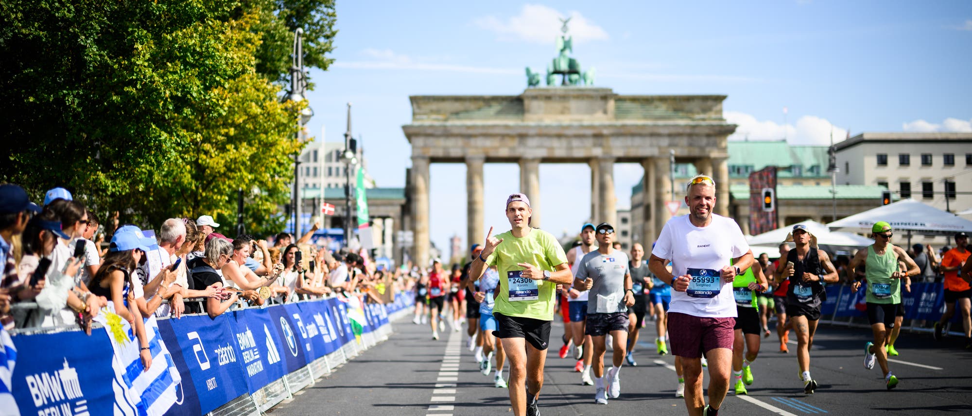 Läufer nehmen an einem Marathon in Berlin teil, während sie auf einer breiten Straße laufen. Im Hintergrund ist das Brandenburger Tor zu sehen und Zuschauer stehen hinter Absperrungen und feuern die Teilnehmer an. Der Himmel ist klar und sonnig, was auf einen schönen Tag hinweist. Die Läufer tragen sportliche Kleidung und Startnummern. Läufer nehmen an einem Marathon in Berlin teil, während sie auf einer breiten Straße laufen. Im Hintergrund ist das Brandenburger Tor zu sehen und Zuschauer stehen hinter Absperrungen und feuern die Teilnehmer an. Der Himmel ist klar und sonnig, was auf einen schönen Tag hinweist. Die Läufer tragen sportliche Kleidung und Startnummern.