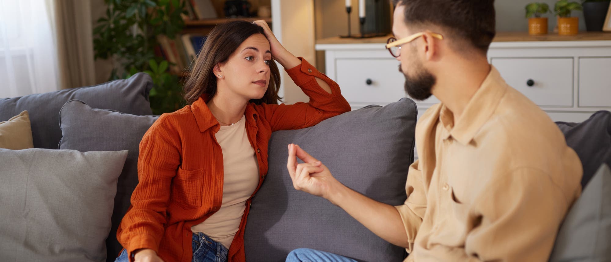 Eine Frau und ein Mann sitzen auf einem Sofa in einem Wohnzimmer und führen ein Gespräch. Die Frau trägt eine orangefarbene Jacke und schaut aufmerksam zu, während der Mann gestikuliert. Im Hintergrund sind Bücherregale, Pflanzen und eine Kommode mit Dekorationen zu sehen. Die Szene vermittelt eine entspannte und häusliche Atmosphäre. Eine Frau und ein Mann sitzen auf einem Sofa in einem Wohnzimmer und führen ein Gespräch. Die Frau trägt eine orangefarbene Jacke und schaut aufmerksam zu, während der Mann gestikuliert. Im Hintergrund sind Bücherregale, Pflanzen und eine Kommode mit Dekorationen zu sehen. Die Szene vermittelt eine entspannte und häusliche Atmosphäre.