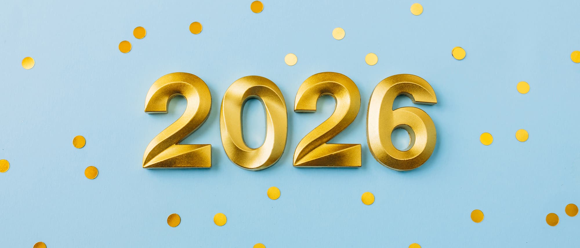 Goldene Zahlen "2026" auf hellblauem Hintergrund, umgeben von verstreuten goldenen Konfetti-Punkten. Die Darstellung vermittelt ein festliches und feierliches Gef&uuml;hl.