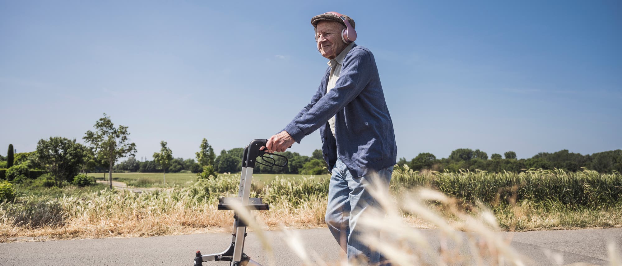 Eine Person geht mit einem Rollator auf einem l&auml;ndlichen Weg entlang. Sie tr&auml;gt Kopfh&ouml;rer und eine Schirmm&uuml;tze. Im Hintergrund sind Felder und B&auml;ume unter einem klaren blauen Himmel zu sehen. Die Szene vermittelt einen ruhigen, sonnigen Tag in der Natur.