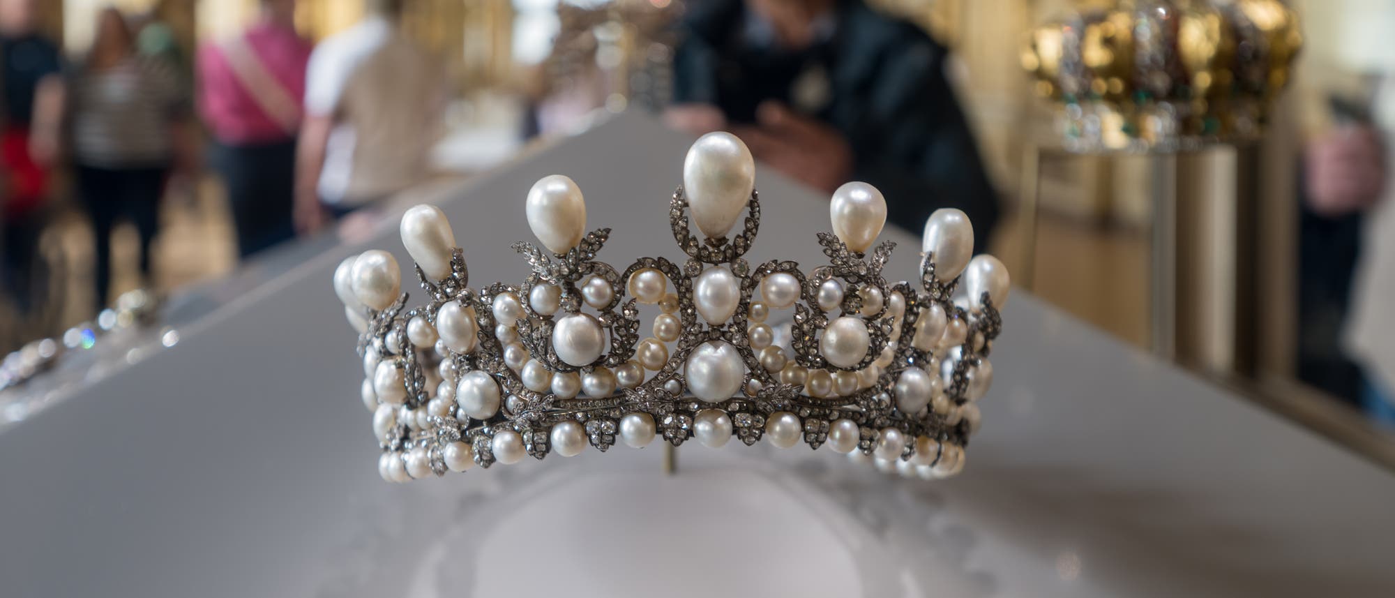 Eine mit Perlen und Diamanten verzierte Tiara wird in einem Museum ausgestellt. Im Hintergrund sind unscharf mehrere Personen zu sehen, die die Ausstellung betrachten. Die Tiara ist auf einem weißen Podest platziert, und ihr Schatten ist deutlich sichtbar. Die Umgebung wirkt elegant und historisch. Eine mit Perlen und Diamanten verzierte Tiara wird in einem Museum ausgestellt. Im Hintergrund sind unscharf mehrere Personen zu sehen, die die Ausstellung betrachten. Die Tiara ist auf einem weißen Podest platziert, und ihr Schatten ist deutlich sichtbar. Die Umgebung wirkt elegant und historisch.