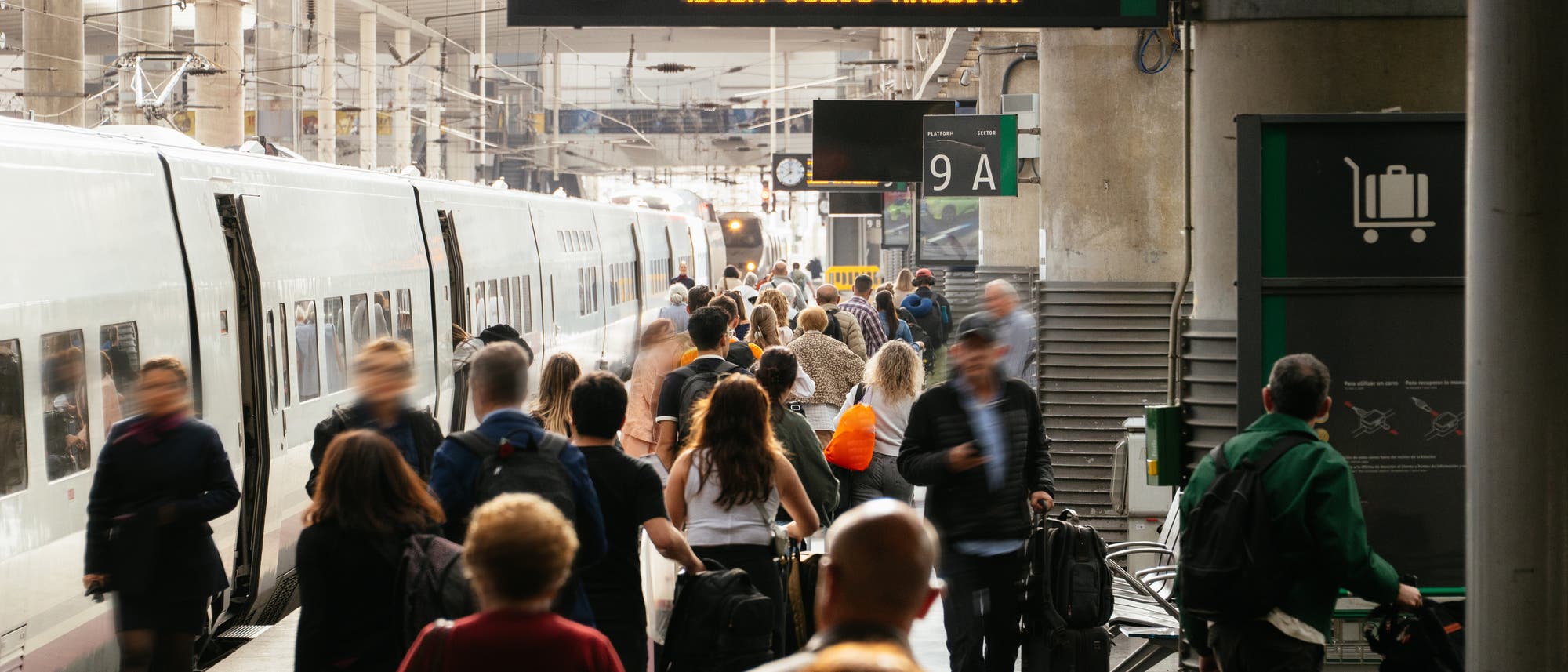Eine belebte Bahnhofsszene mit vielen Menschen, die auf einem Bahnsteig gehen. Ein Zug steht auf der linken Seite, während eine Anzeigetafel mit der Aufschrift „Platform 9“ und einer Uhr darüber sichtbar ist. Die Menschen tragen Gepäck und scheinen in Eile zu sein. Im Hintergrund sind weitere Züge und Bahnsteige zu sehen. Eine belebte Bahnhofsszene mit vielen Menschen, die auf einem Bahnsteig gehen. Ein Zug steht auf der linken Seite, während eine Anzeigetafel mit der Aufschrift „Platform 9“ und einer Uhr darüber sichtbar ist. Die Menschen tragen Gepäck und scheinen in Eile zu sein. Im Hintergrund sind weitere Züge und Bahnsteige zu sehen.