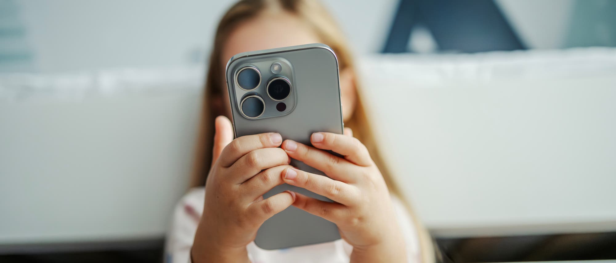 Ein Kind h&auml;lt ein modernes Smartphone mit beiden H&auml;nden und betrachtet den Bildschirm. Das Kind tr&auml;gt ein Oberteil mit Herzmustern. Der Hintergrund ist unscharf und zeigt helle Farben.