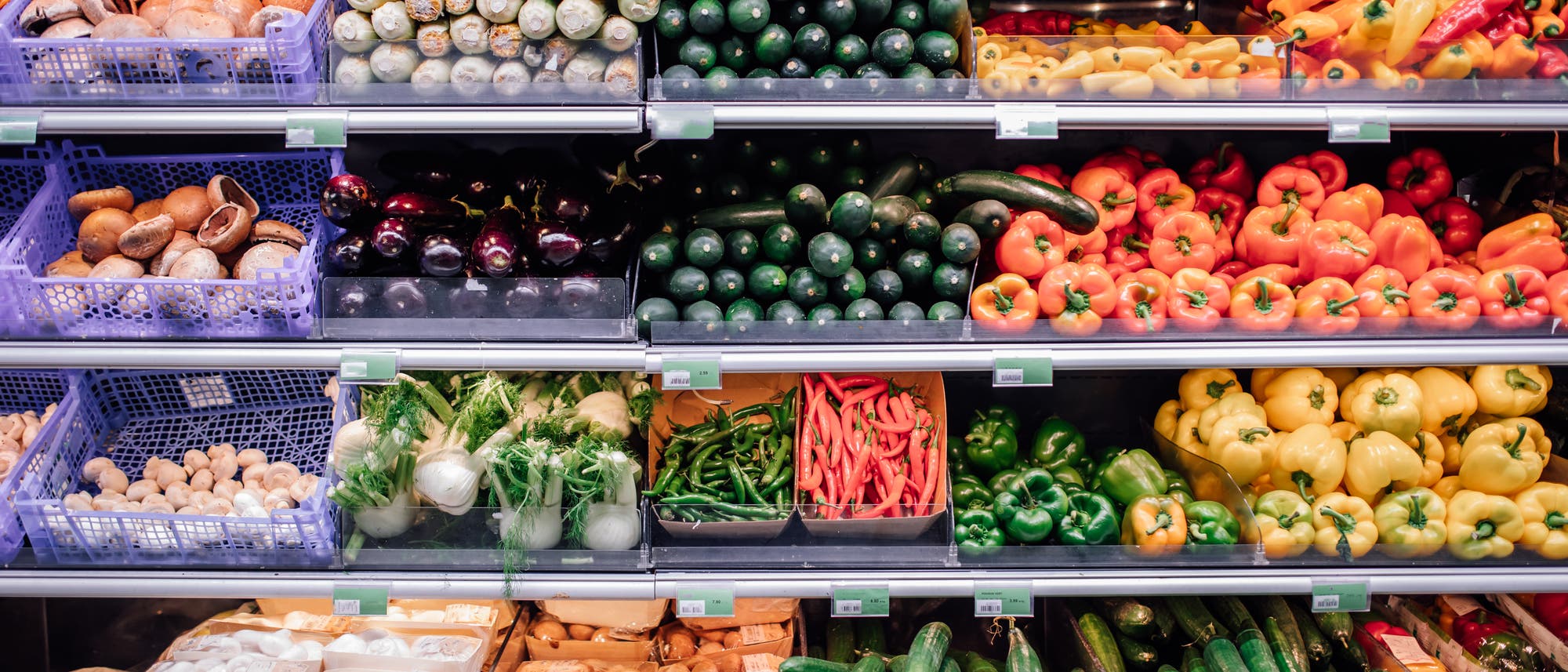 Ein Supermarktregal mit einer Vielzahl von frischem Gemüse. Oben links befinden sich Pilze in blauen Kisten, daneben Zucchini und Gurken. In der Mitte sind Auberginen, Paprika in verschiedenen Farben und grüne Chilischoten zu sehen. Unten sind Fenchel, weitere Pilze und Gurken angeordnet. Die Produkte sind ordentlich in Reihen gestapelt und bieten eine bunte Auswahl an frischen Lebensmitteln. Ein Supermarktregal mit einer Vielzahl von frischem Gemüse. Oben links befinden sich Pilze in blauen Kisten, daneben Zucchini und Gurken. In der Mitte sind Auberginen, Paprika in verschiedenen Farben und grüne Chilischoten zu sehen. Unten sind Fenchel, weitere Pilze und Gurken angeordnet. Die Produkte sind ordentlich in Reihen gestapelt und bieten eine bunte Auswahl an frischen Lebensmitteln.