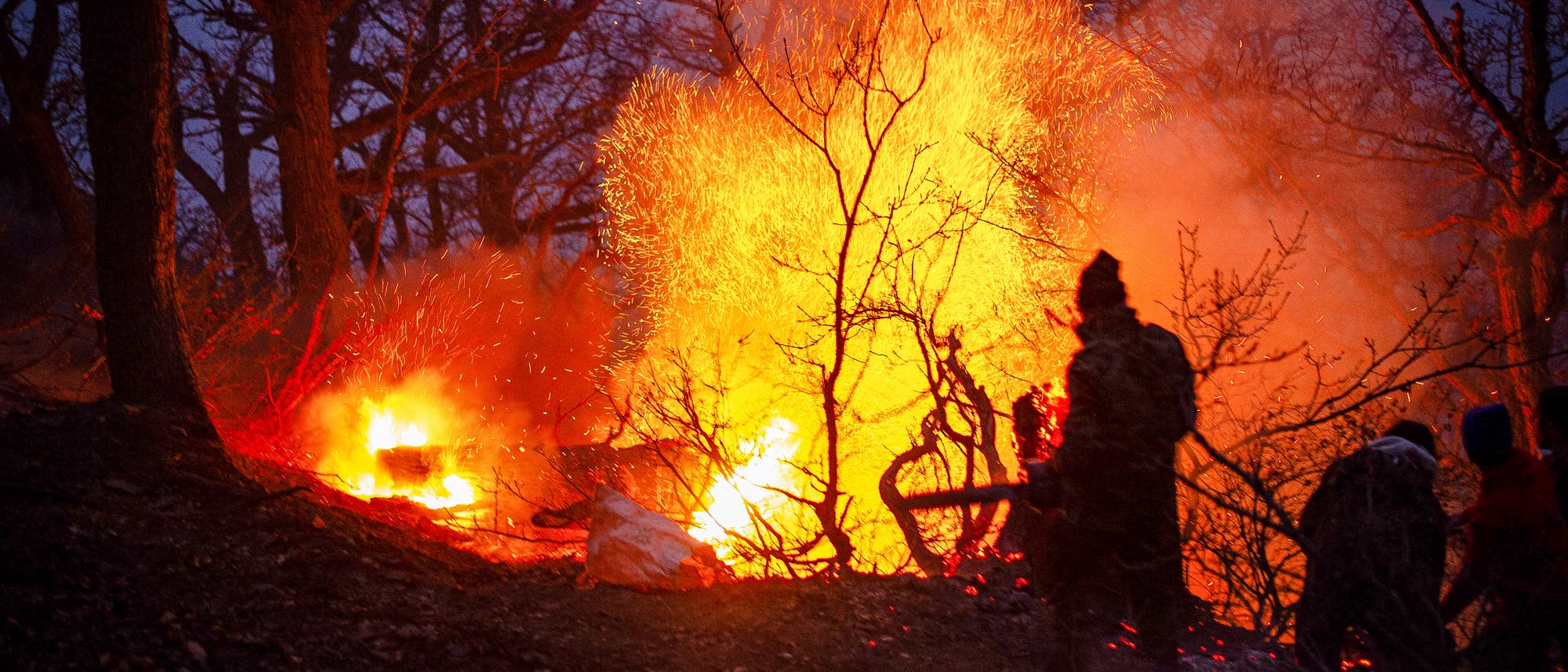 Eine Gruppe von Menschen steht in einem Wald bei Nacht, w&auml;hrend ein gro&szlig;es Feuer lodert. Die Flammen leuchten hell und werfen Funken in die Luft, w&auml;hrend Rauch aufsteigt. Die Silhouetten der Personen sind vor dem Hintergrund des Feuers sichtbar, umgeben von kahlen B&auml;umen. Die Szene vermittelt eine dramatische und intensive Atmosph&auml;re.