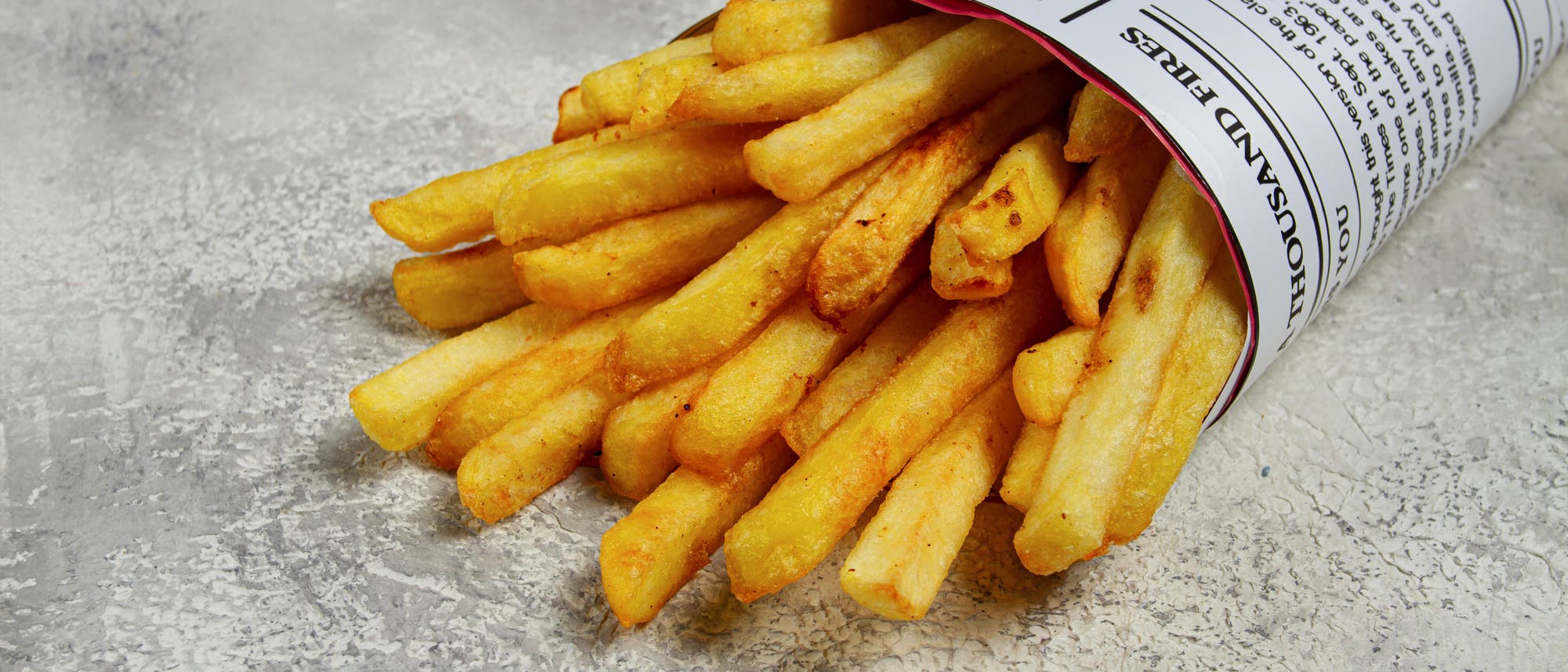 Eine Portion Pommes frites, die in einem Papierkegel serviert werden. Der Kegel besteht aus bedrucktem Papier mit Text. Die Pommes sind goldbraun und knusprig, auf einer grauen, strukturierten Oberfläche platziert. Eine Portion Pommes frites, die in einem Papierkegel serviert werden. Der Kegel besteht aus bedrucktem Papier mit Text. Die Pommes sind goldbraun und knusprig, auf einer grauen, strukturierten Oberfläche platziert.