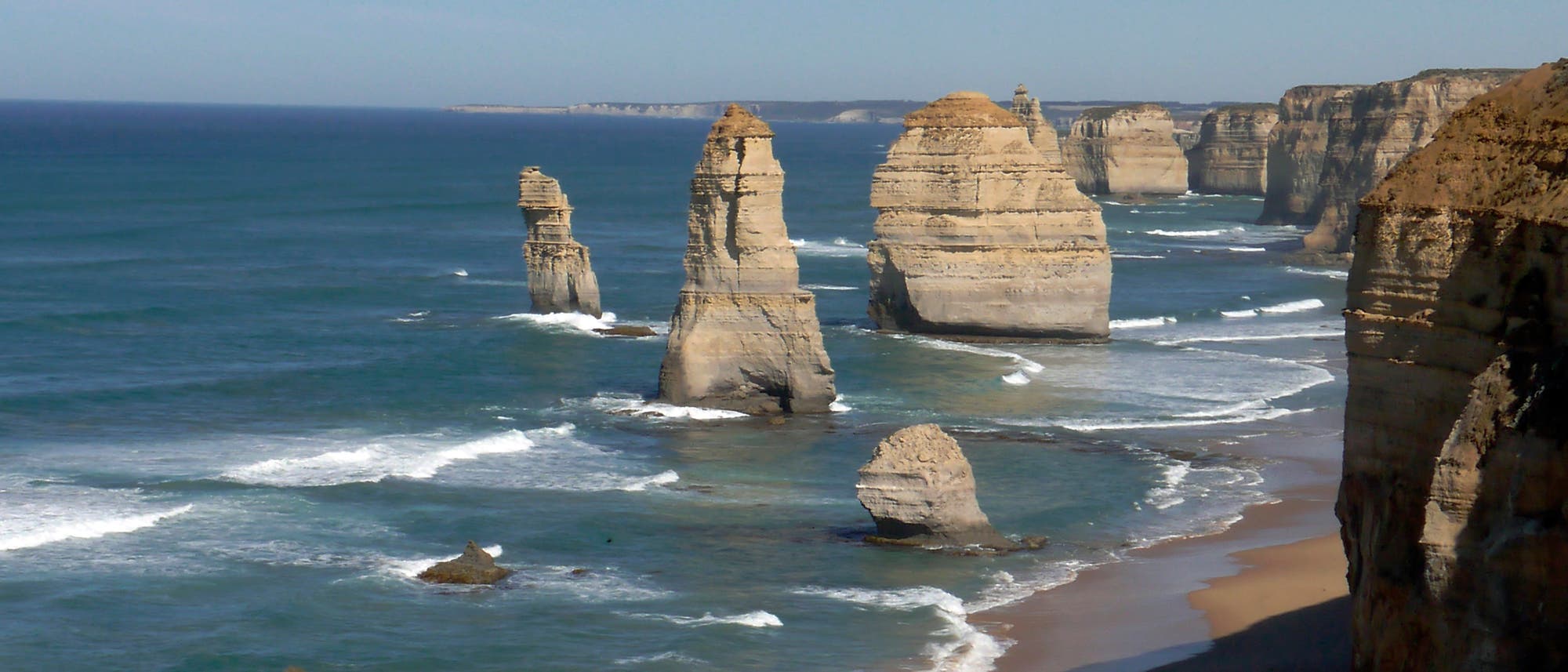K&uuml;stenlandschaft mit mehreren markanten Kalksteinfelsen, die aus dem Meer ragen. Die Felsen sind von Wellen umsp&uuml;lt und stehen vor einer steilen Klippe. Der Himmel ist klar und das Meer erstreckt sich bis zum Horizont.