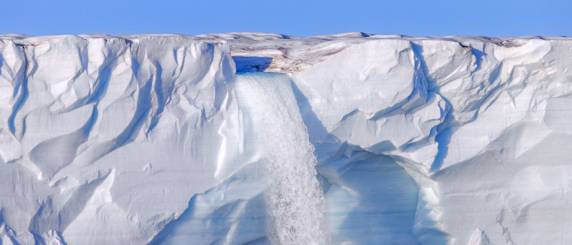 Ein gro&szlig;er Gletscher mit einer Wasserfallkaskade, die von der Eiskante in das darunterliegende Meer st&uuml;rzt. Die Oberfl&auml;che des Gletschers ist uneben und von Rissen durchzogen. Der Himmel ist klar und blau, was die k&uuml;hle, arktische Umgebung betont.