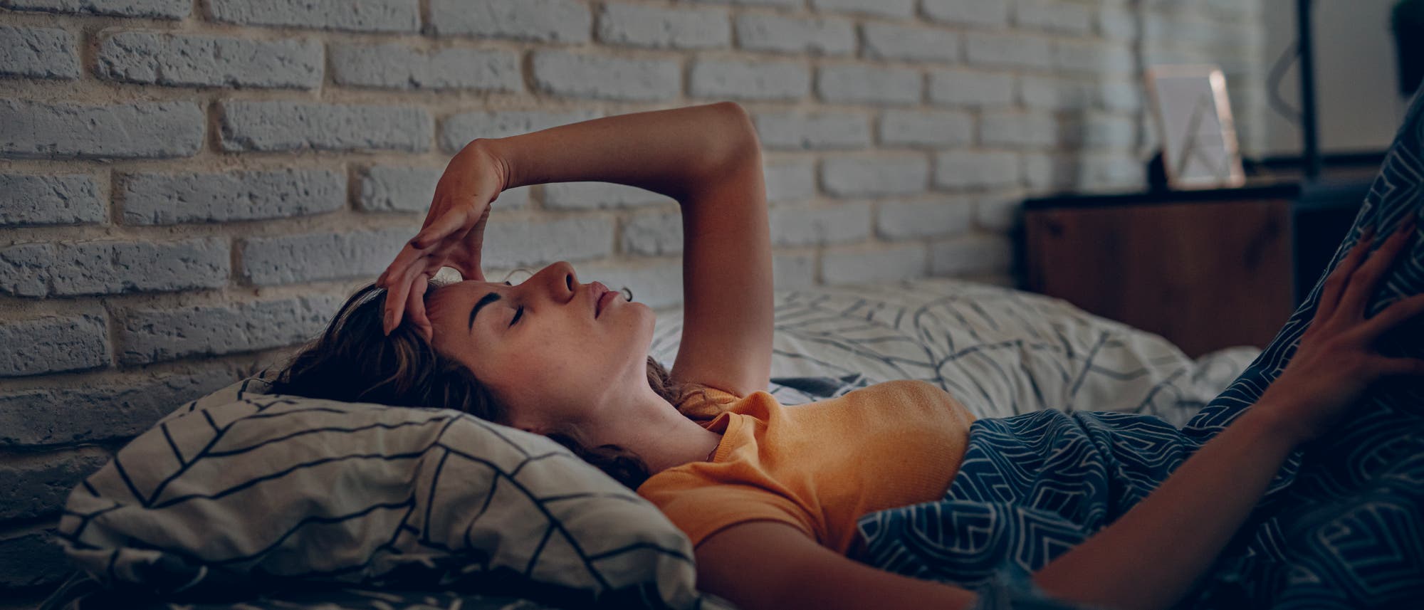 Eine Person liegt in einem Bett, umgeben von gemusterten Kissen und einer Decke. Sie trägt ein gelbes Oberteil und hat eine Hand auf der Stirn, als ob sie nachdenkt oder sich ausruht. Im Hintergrund ist eine weiße Ziegelwand zu sehen. Die Szene vermittelt eine ruhige und entspannte Atmosphäre. Eine Person liegt in einem Bett, umgeben von gemusterten Kissen und einer Decke. Sie trägt ein gelbes Oberteil und hat eine Hand auf der Stirn, als ob sie nachdenkt oder sich ausruht. Im Hintergrund ist eine weiße Ziegelwand zu sehen. Die Szene vermittelt eine ruhige und entspannte Atmosphäre.