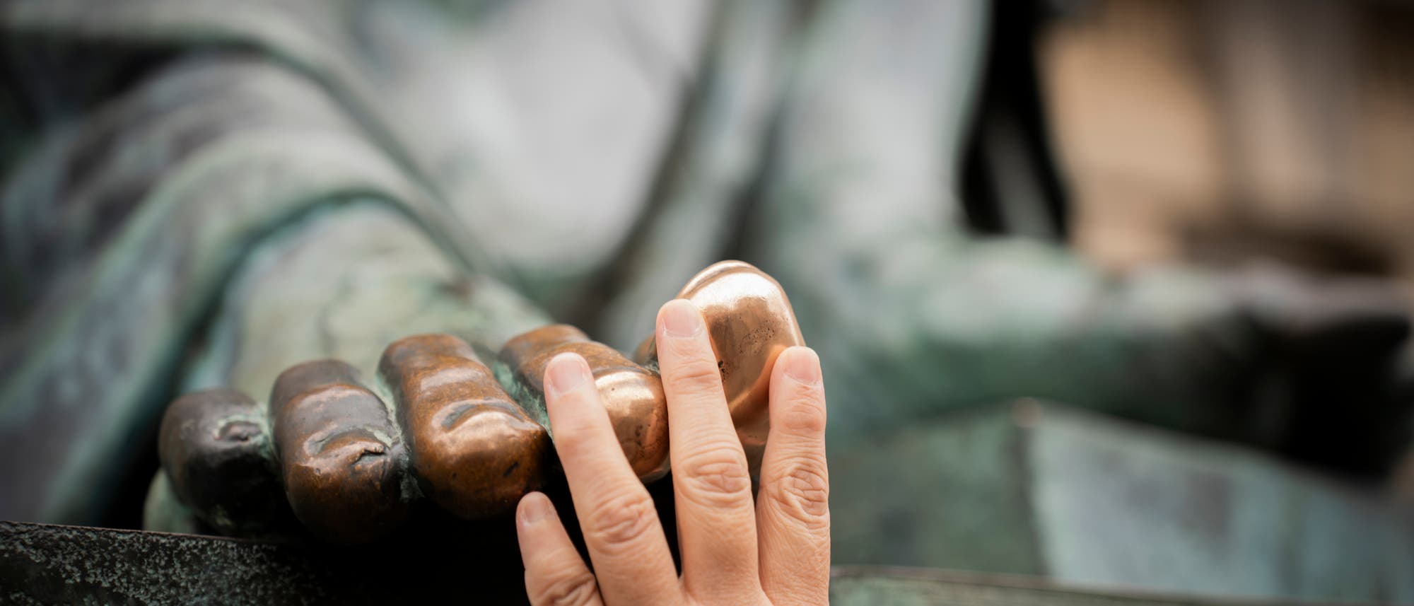 Eine Hand ber&uuml;hrt die gl&auml;nzende, abgenutzte Oberfl&auml;che der Finger einer gro&szlig;en Bronzestatue. Die Statue ist teilweise sichtbar und zeigt eine ausgestreckte Hand. Die Ber&uuml;hrung hat die Patina der Statue an dieser Stelle abgetragen, was auf h&auml;ufigen Kontakt hinweist. Im Hintergrund sind verschwommene Details der Statue zu erkennen.