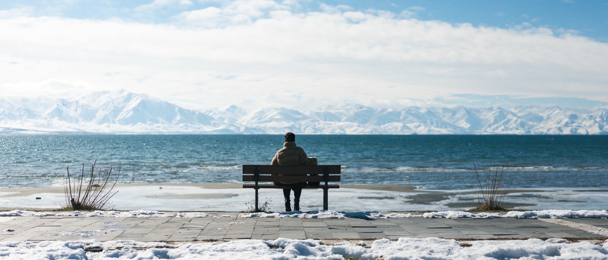 Eine Person sitzt auf einer Bank und blickt auf einen ruhigen See, der von schneebedeckten Bergen umgeben ist. Der Boden ist teilweise mit Schnee bedeckt, und der Himmel ist klar mit einigen Wolken. Die Szene vermittelt eine ruhige und friedliche Winterlandschaft. Eine Person sitzt auf einer Bank und blickt auf einen ruhigen See, der von schneebedeckten Bergen umgeben ist. Der Boden ist teilweise mit Schnee bedeckt, und der Himmel ist klar mit einigen Wolken. Die Szene vermittelt eine ruhige und friedliche Winterlandschaft.