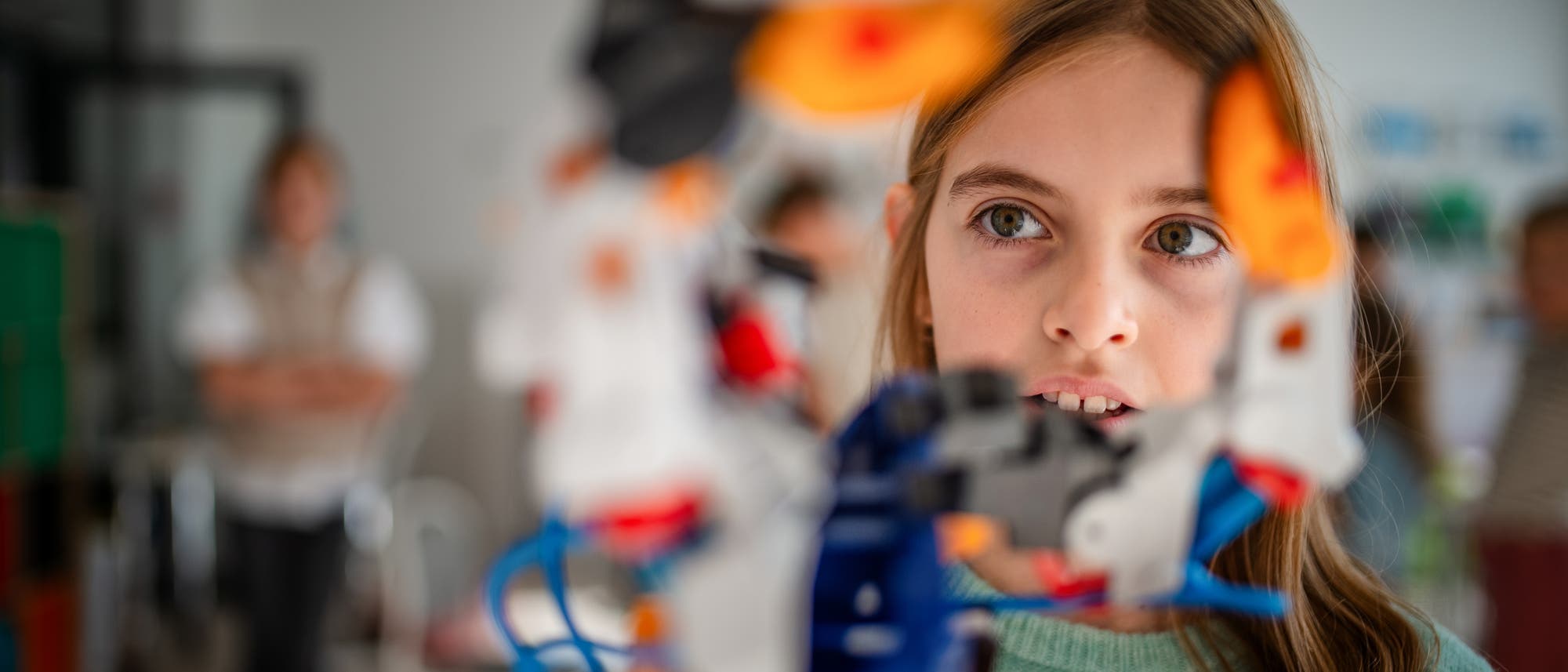 Ein Kind betrachtet konzentriert ein buntes Roboter-Modell in einem Klassenzimmer. Im Hintergrund sind unscharf weitere Personen und M&ouml;bel zu erkennen. Das Bild vermittelt eine Lernumgebung, in der digitale Technologie und Kreativit&auml;t im Vordergrund stehen.
