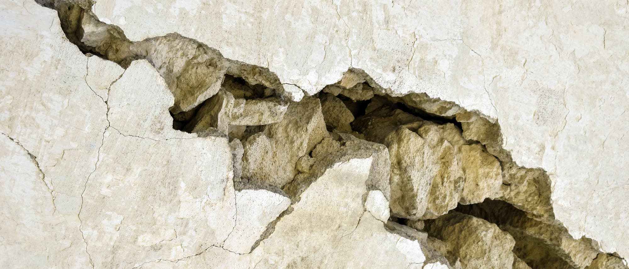 Riss in einer beigen Steinwand, der unregelm&auml;&szlig;ige Bruchlinien und freiliegende Gesteinsbrocken zeigt. Die Oberfl&auml;che weist feine Risse und Texturen auf, die auf nat&uuml;rliche Erosion oder strukturelle Sch&auml;den hindeuten.