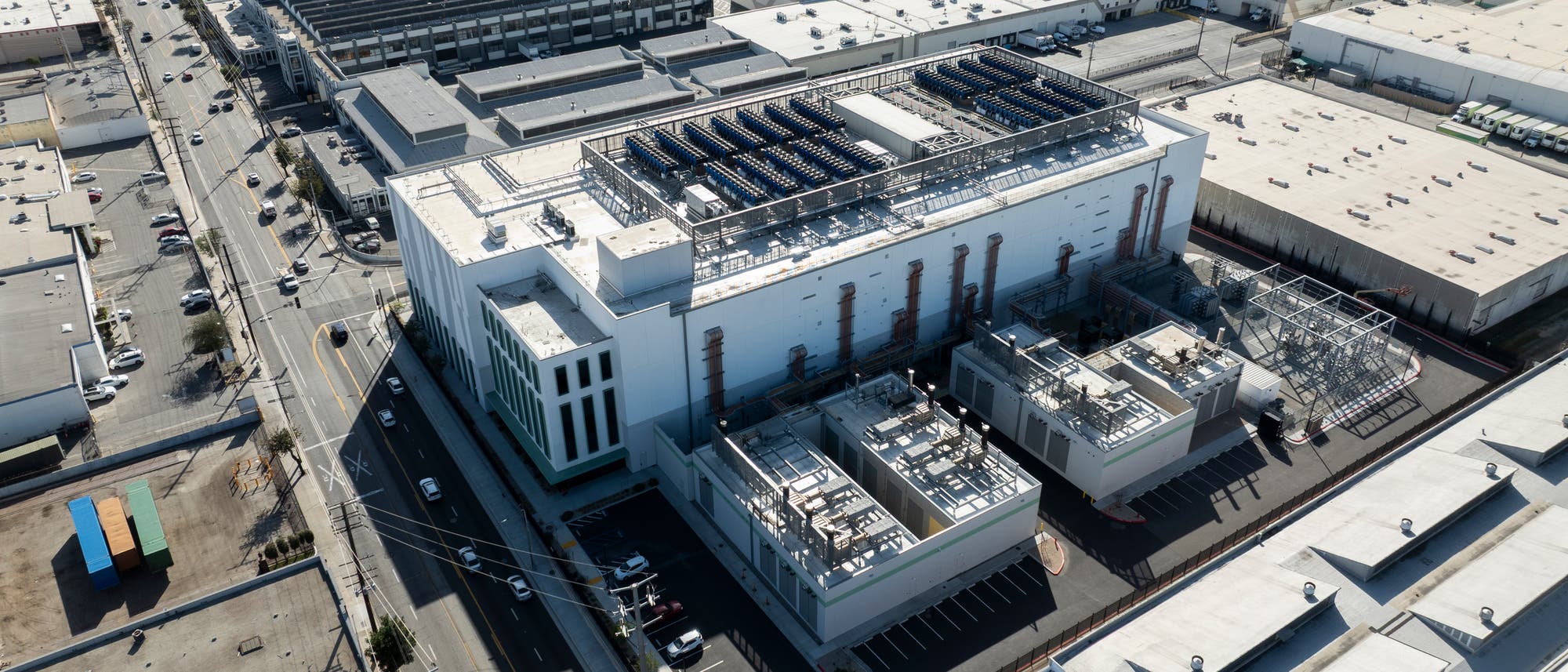 Luftaufnahme eines gro&szlig;en Industriegeb&auml;udes in einem st&auml;dtischen Gebiet. Das Geb&auml;ude hat eine wei&szlig;e Fassade und ist mit mehreren Solarpaneelen auf dem Dach ausgestattet. Um das Geb&auml;ude herum befinden sich Parkpl&auml;tze und kleinere technische Strukturen. Die Umgebung zeigt weitere Industriegeb&auml;ude und Stra&szlig;en mit vereinzeltem Verkehr.