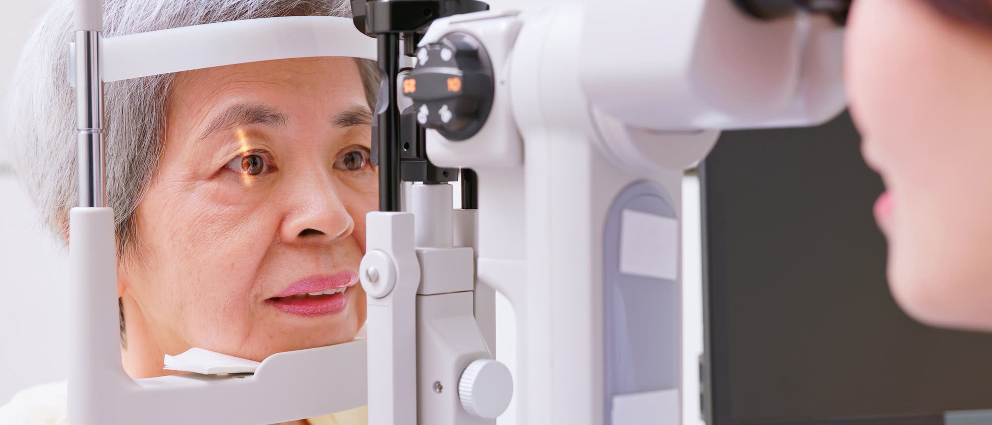 Eine Person sitzt vor einem ophthalmologischen Ger&auml;t und unterzieht sich einer Augenuntersuchung. Die Person blickt konzentriert in das Ger&auml;t, w&auml;hrend ein medizinisches Fachpersonal die Untersuchung durchf&uuml;hrt. Das Bild zeigt die klinische Umgebung und die Verwendung moderner Technologie zur Augenuntersuchung.