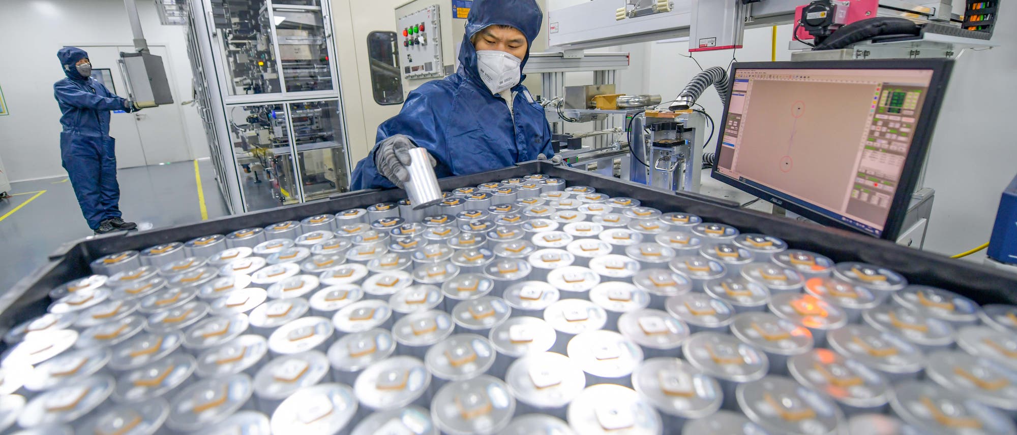 Eine Person in Schutzkleidung und Maske arbeitet in einem Labor an einer gro&szlig;en Anzahl von zylindrischen Batteriezellen, die auf einem Tisch angeordnet sind. Im Hintergrund ist ein weiterer Mitarbeiter an einer Maschine zu sehen. Ein Computerbildschirm zeigt technische Daten an. Die Szene vermittelt den Eindruck einer kontrollierten Umgebung zur Herstellung oder Pr&uuml;fung von Batterien.