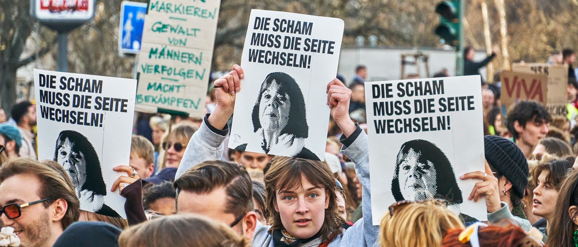 Eine Menschenmenge bei einer Demonstration hält Schilder hoch. Auf den Schildern steht: „Die Scham muss die Seite wechseln!“ und zeigen das Bild einer Frau. Im Hintergrund sind weitere Schilder mit Botschaften gegen Gewalt zu sehen. Die Szene findet im Freien statt, umgeben von Bäumen und Verkehrsschildern. Eine Menschenmenge bei einer Demonstration hält Schilder hoch. Auf den Schildern steht: „Die Scham muss die Seite wechseln!“ und zeigen das Bild einer Frau. Im Hintergrund sind weitere Schilder mit Botschaften gegen Gewalt zu sehen. Die Szene findet im Freien statt, umgeben von Bäumen und Verkehrsschildern.