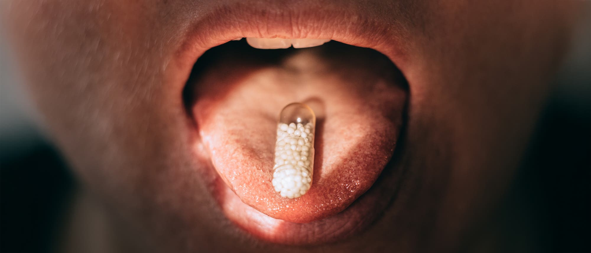 Eine Nahaufnahme eines geöffneten Mundes mit einer transparenten Kapsel auf der Zunge. Die Kapsel enthält kleine weiße Kügelchen. Das Bild zeigt den Moment des Einnehmens eines Medikaments. Eine Nahaufnahme eines geöffneten Mundes mit einer transparenten Kapsel auf der Zunge. Die Kapsel enthält kleine weiße Kügelchen. Das Bild zeigt den Moment des Einnehmens eines Medikaments.