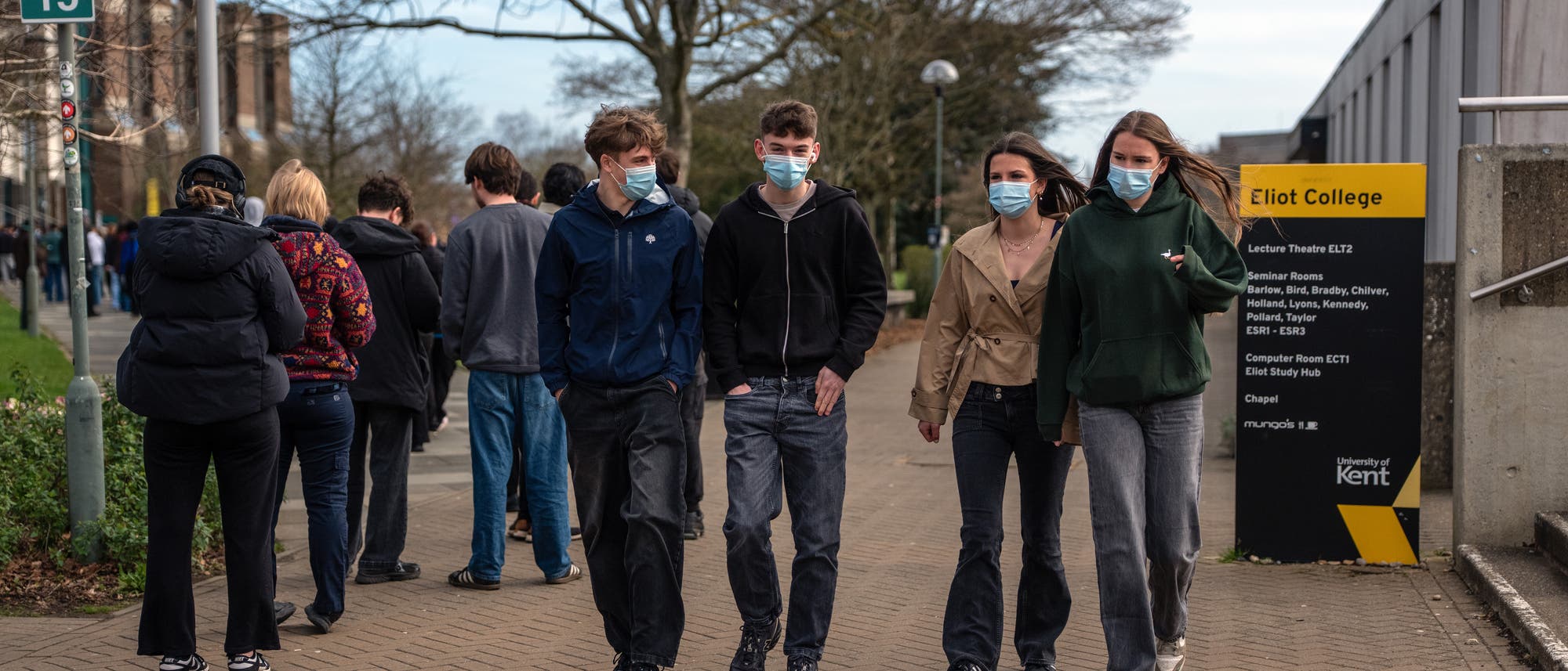 Eine Gruppe von vier jungen Menschen, die Gesichtsmasken tragen, geht auf einem Campusweg. Im Hintergrund sind weitere Personen zu sehen, die in einer Schlange anstehen. Rechts im Bild steht ein Schild mit der Aufschrift &bdquo;Eliot College&ldquo; und einer Liste von R&auml;umen und Einrichtungen der University of Kent. 