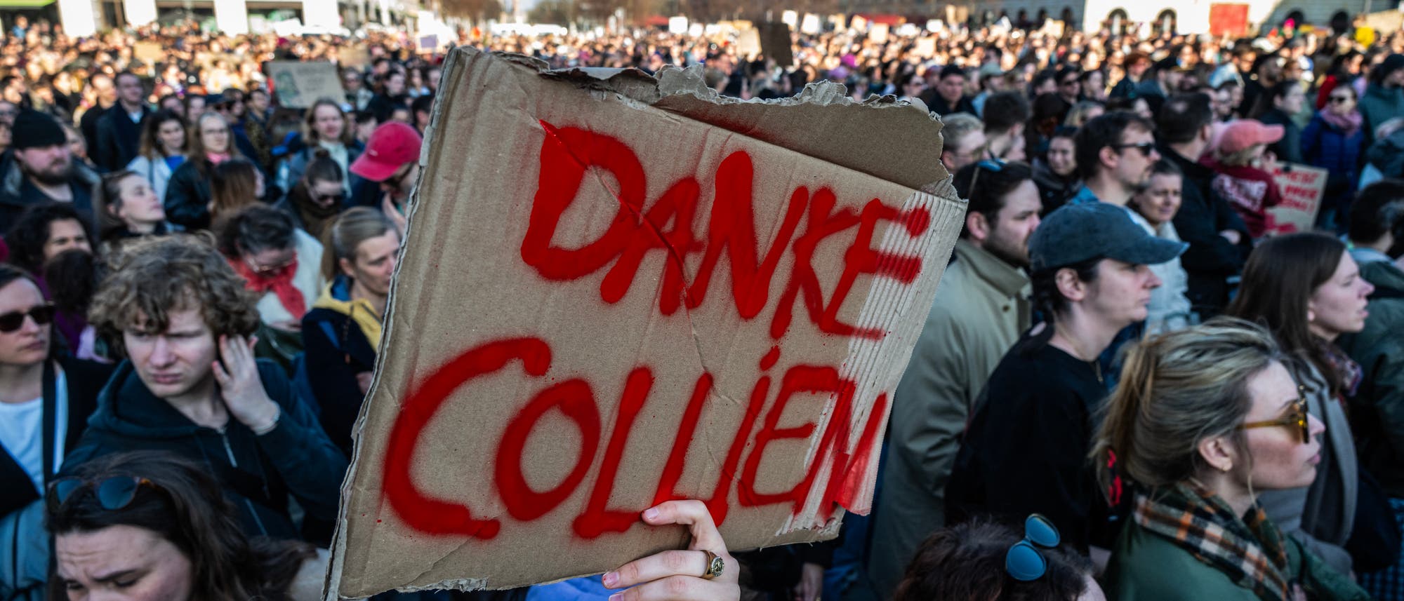Eine gro&szlig;e Menschenmenge versammelt sich bei einer Demonstration im Freien. Im Vordergrund h&auml;lt jemand ein handgemaltes Schild aus Pappe mit der Aufschrift &bdquo;DANKE COLLIEN&ldquo;. Die Menschen wirken aufmerksam und engagiert. Im Hintergrund sind moderne Geb&auml;ude zu sehen. Die Szene vermittelt eine Atmosph&auml;re von Gemeinschaft und Engagement.