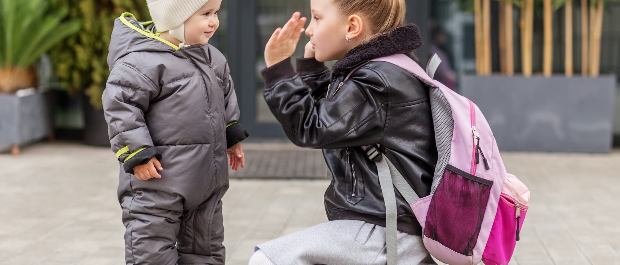 Zwei Kinder stehen drau&szlig;en vor einem Geb&auml;ude. Das j&uuml;ngere Kind tr&auml;gt einen grauen Schneeanzug und eine cremefarbene M&uuml;tze. Das &auml;ltere Kind, mit einem rosa Rucksack, kniet und gestikuliert freundlich. Im Hintergrund sind Pflanzen und eine Glast&uuml;r zu sehen. Die Szene wirkt fr&ouml;hlich und zeigt eine Interaktion zwischen den Kindern.