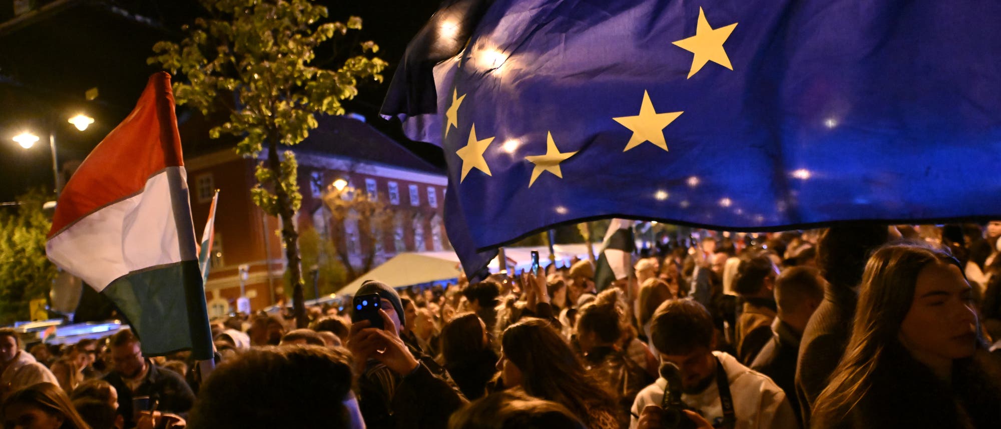 Eine Menschenmenge versammelt sich nachts auf einer Stra&szlig;e. Im Vordergrund wehen eine EU-Flagge und eine ungarische Flagge. Die Szene ist belebt, mit vielen Menschen, die Fotos machen und die Atmosph&auml;re genie&szlig;en. Im Hintergrund sind beleuchtete Geb&auml;ude und B&auml;ume zu sehen.
