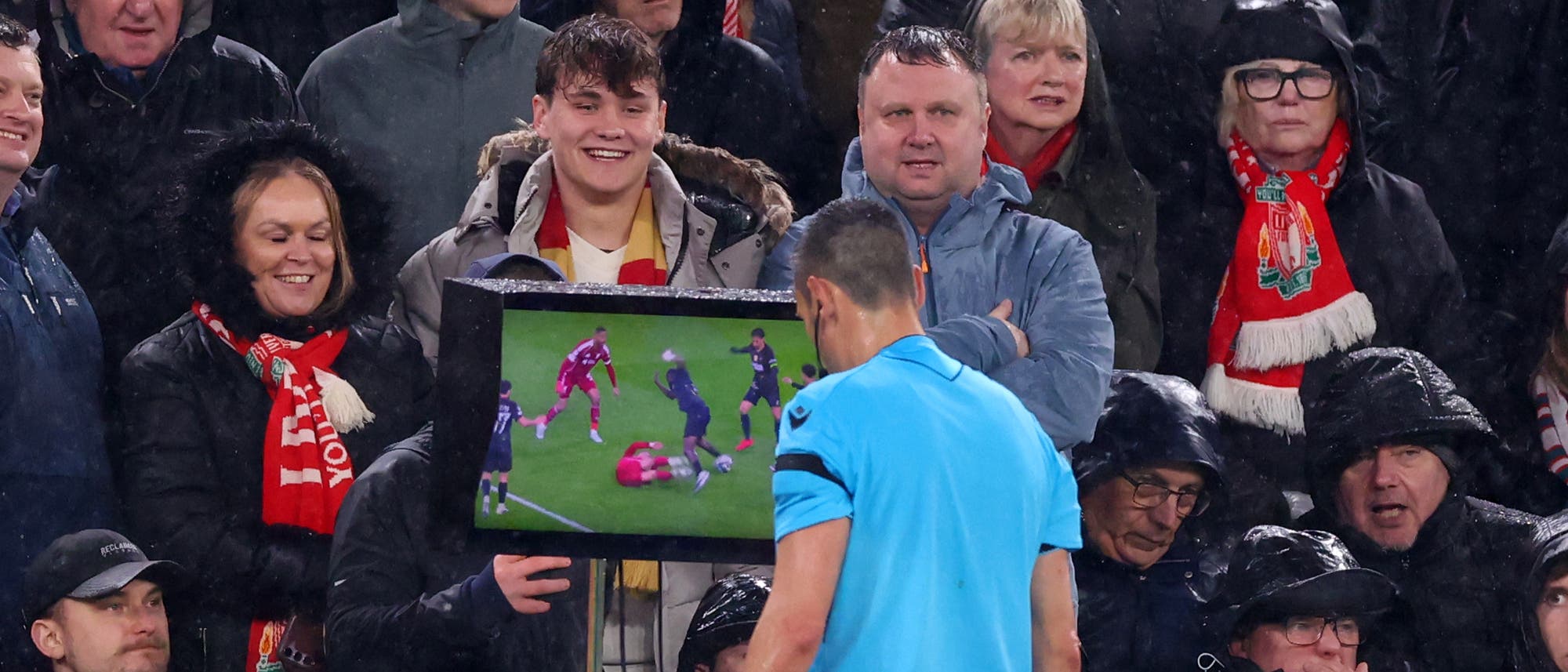 Ein Schiedsrichter in einem blauen Trikot betrachtet einen Bildschirm am Spielfeldrand, w&auml;hrend eine Gruppe von Fu&szlig;ballfans im Hintergrund zuschaut. Der Bildschirm zeigt eine Spielsituation mit mehreren Spielern auf dem Fu&szlig;ballfeld. Die Fans tragen Regenkleidung und Schals in den Farben eines Fu&szlig;ballvereins. Die Szene findet bei Regen statt, was an den nassen Kleidern der Zuschauer erkennbar ist.