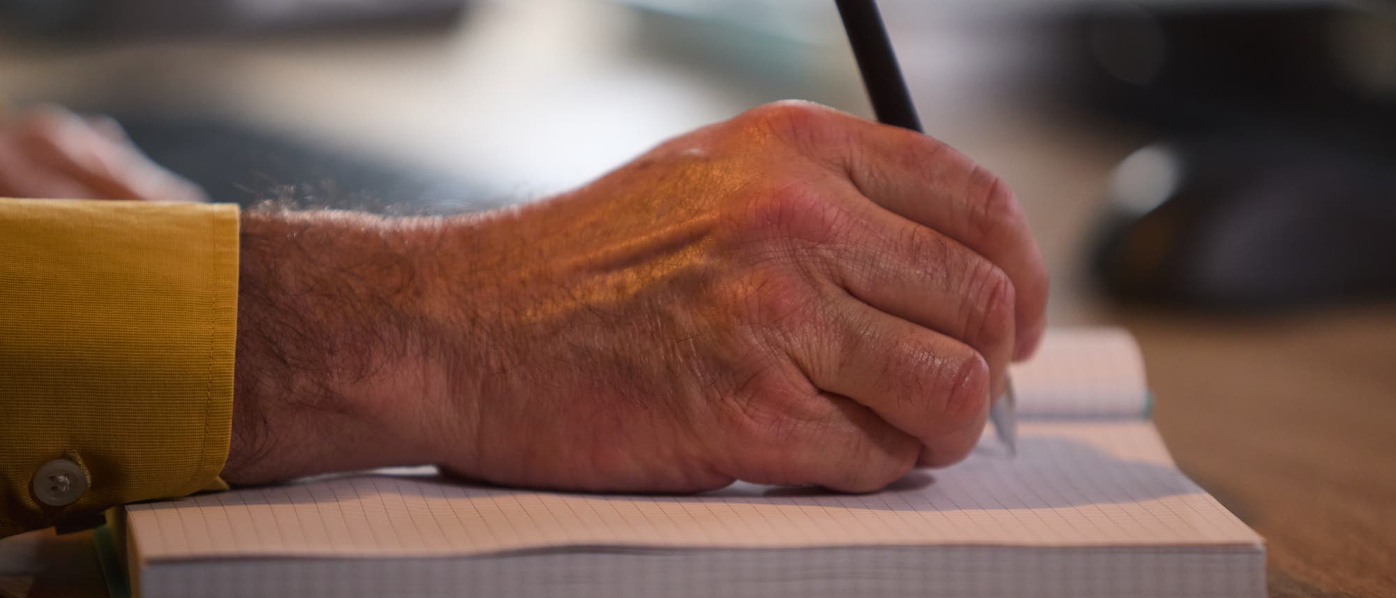 Eine Person schreibt mit einem schwarzen Stift in ein Notizbuch mit kariertem Papier. Die Person trägt ein gelbes Hemd. Eine Person schreibt mit einem schwarzen Stift in ein Notizbuch mit kariertem Papier. Die Person trägt ein gelbes Hemd.