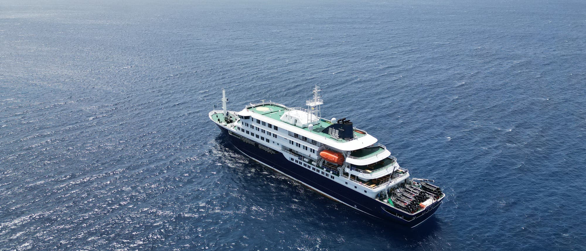 Ein gro&szlig;es Kreuzfahrtschiff f&auml;hrt auf offenem Meer. Das Schiff hat mehrere Decks, darunter ein gr&uuml;nes Oberdeck. Rettungsboote sind an der Seite sichtbar. Der Himmel ist leicht bew&ouml;lkt, und das Wasser ist ruhig.